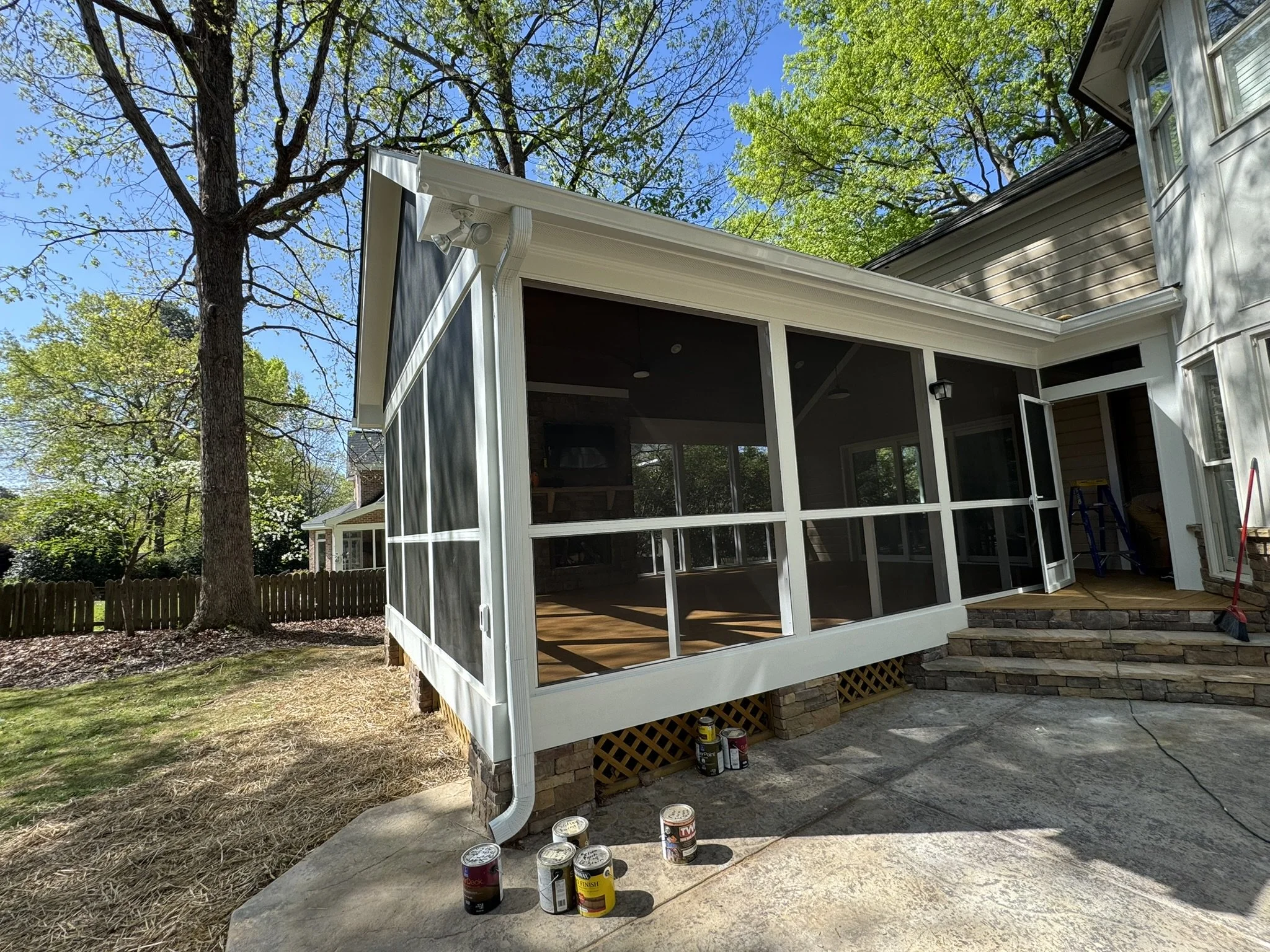 A screened porch attached to a house with a brick foundation, surrounded by trees with green leaves, and a pile of paint cans on the concrete patio below.