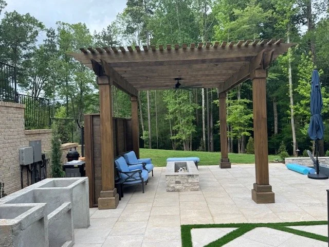 Outdoor patio with a wooden pergola, seating area, fire pit, and patio furniture, surrounded by trees.