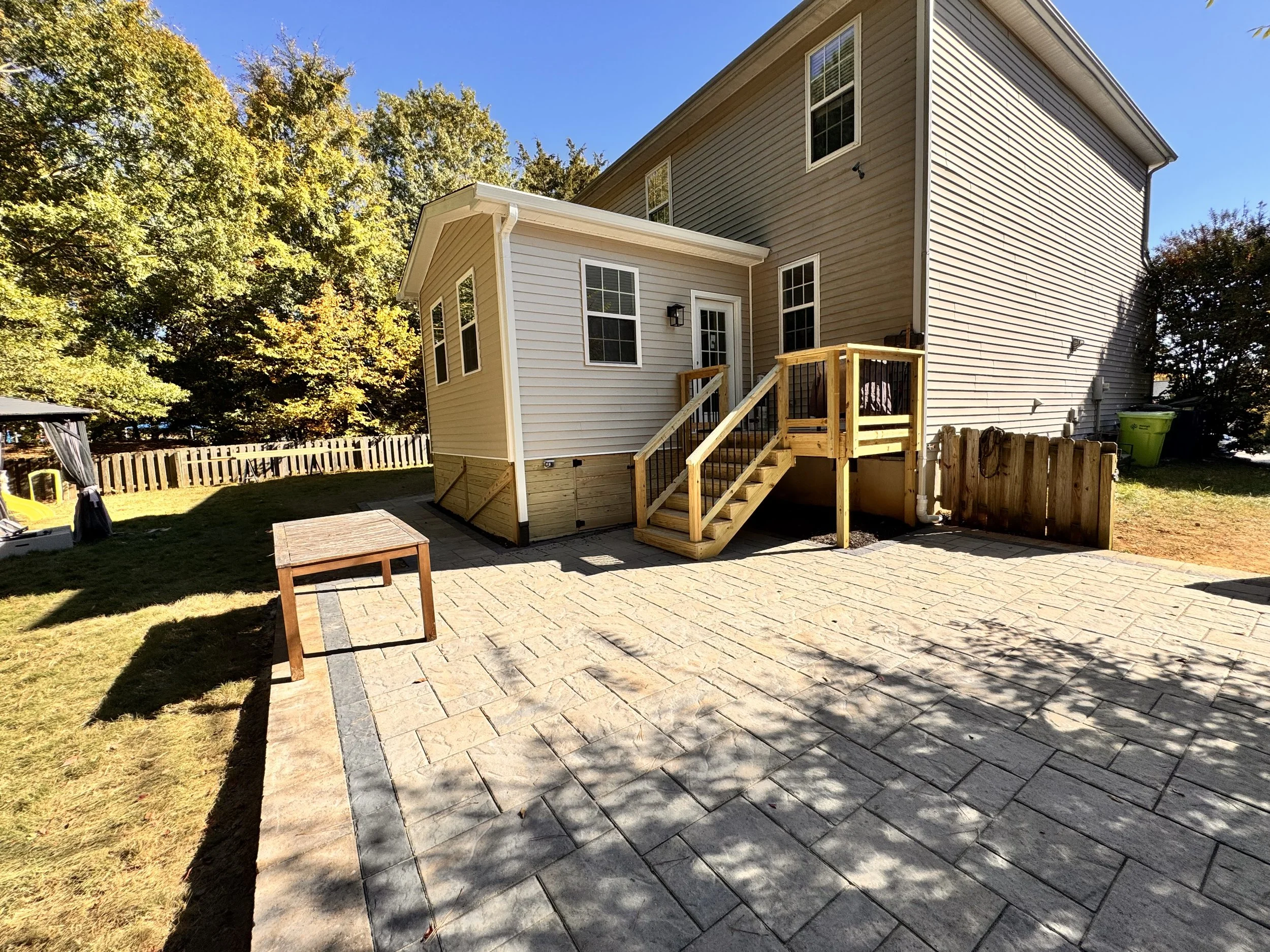 Backyard view of a two-story house with a small wooden staircase leading up to a deck door. There is a brick patio area with a small table, wood fencing, and green trash bins. Trees with green and yellow leaves are in the background under a blue sky.