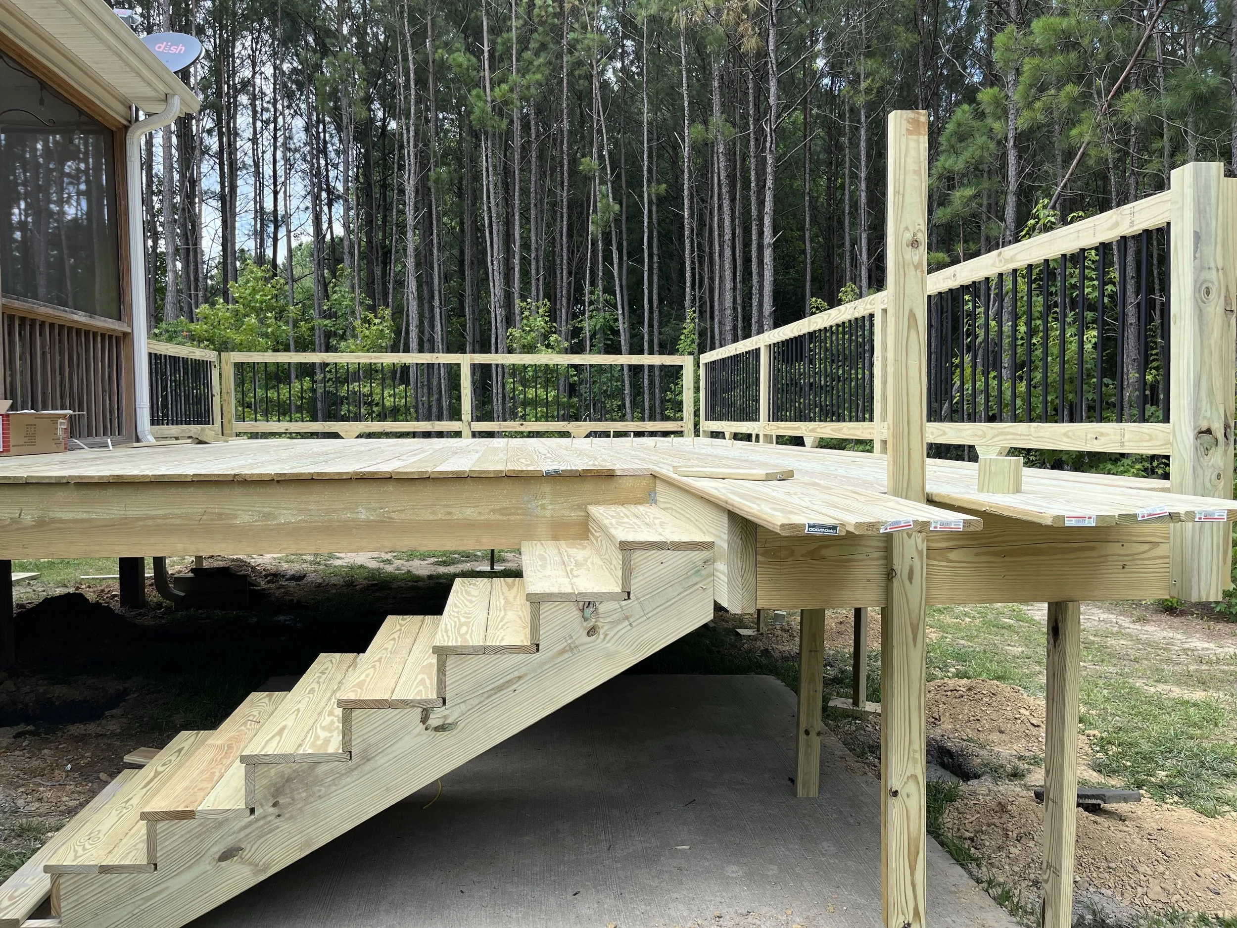A partially constructed wooden deck with railing, stairs, and a view of a trees in the background.