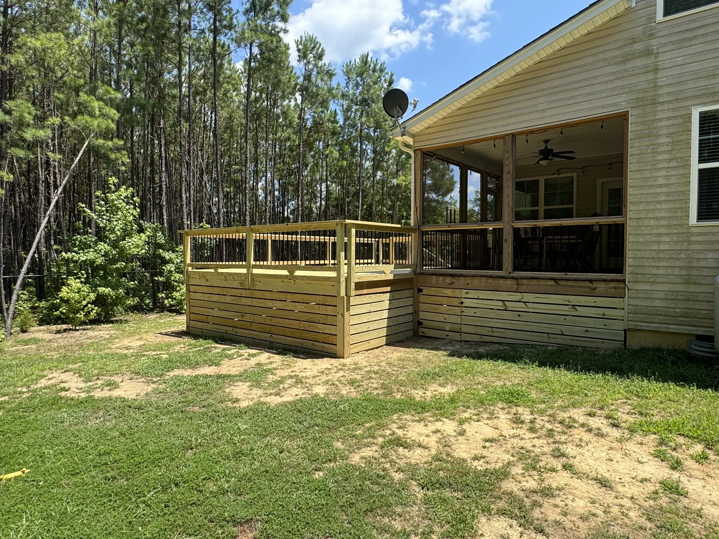Back patio of a house with a screened porch, wooden deck, satellite dish, and string lights, surrounded by a forest of tall pine trees under a partly cloudy sky.