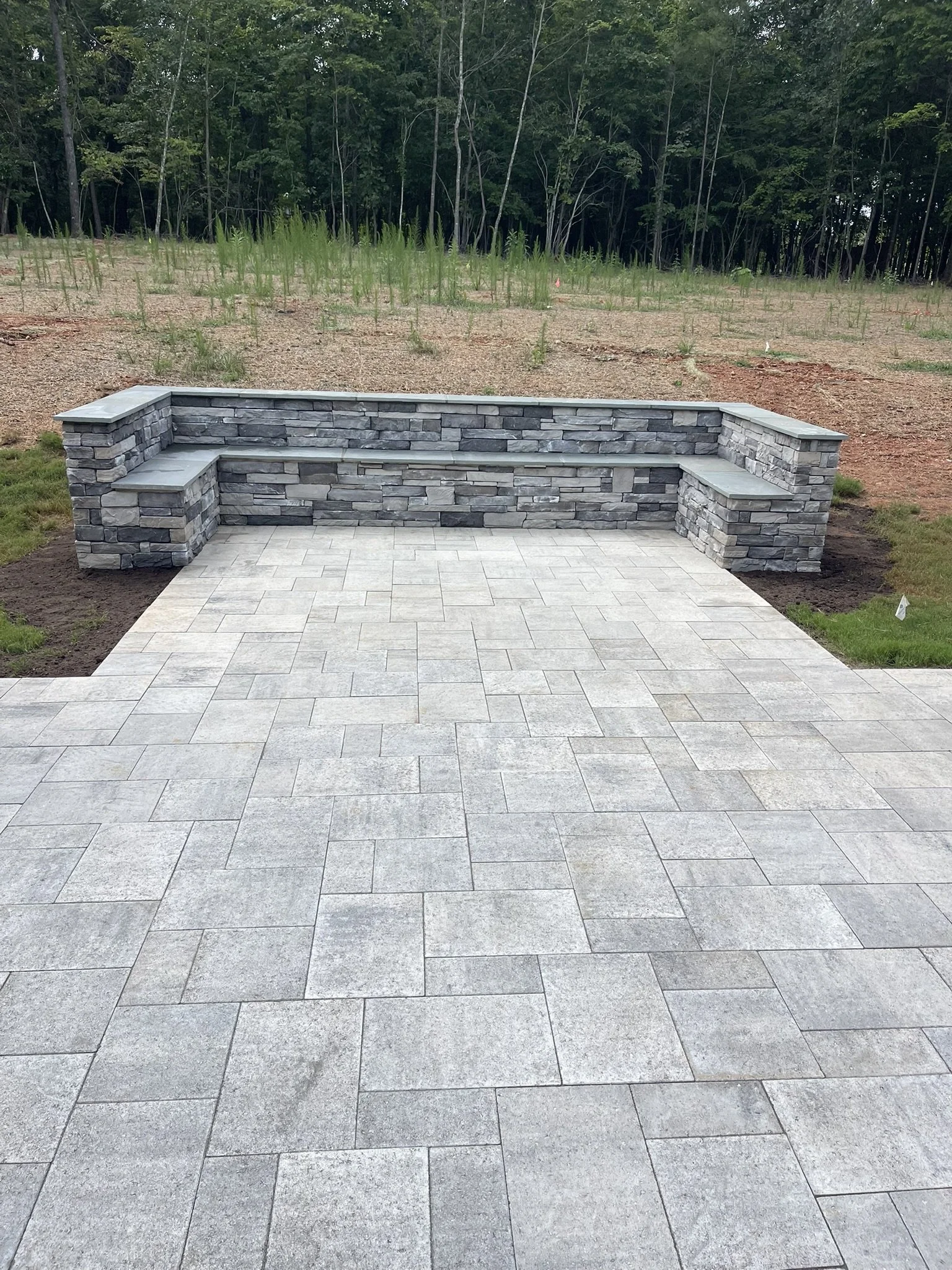 Stone and brick outdoor bench with a tiled patio in front, green grass and dirt area, and a dense forest background.