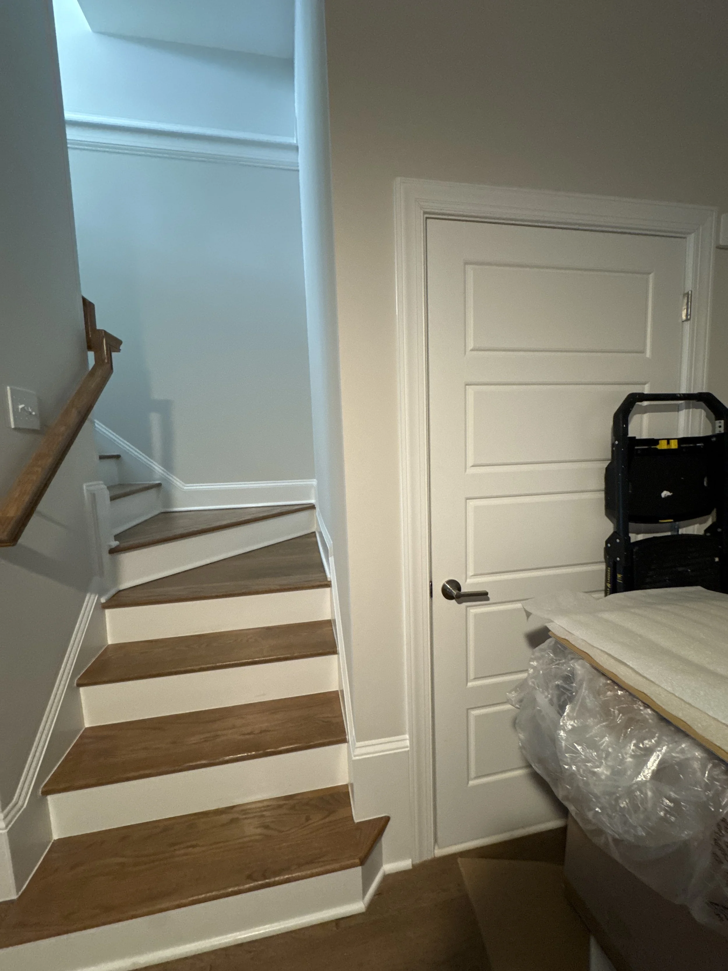 Wooden staircase with dark brown steps and white risers, near a white closed door and a black step ladder in a room under renovation.
