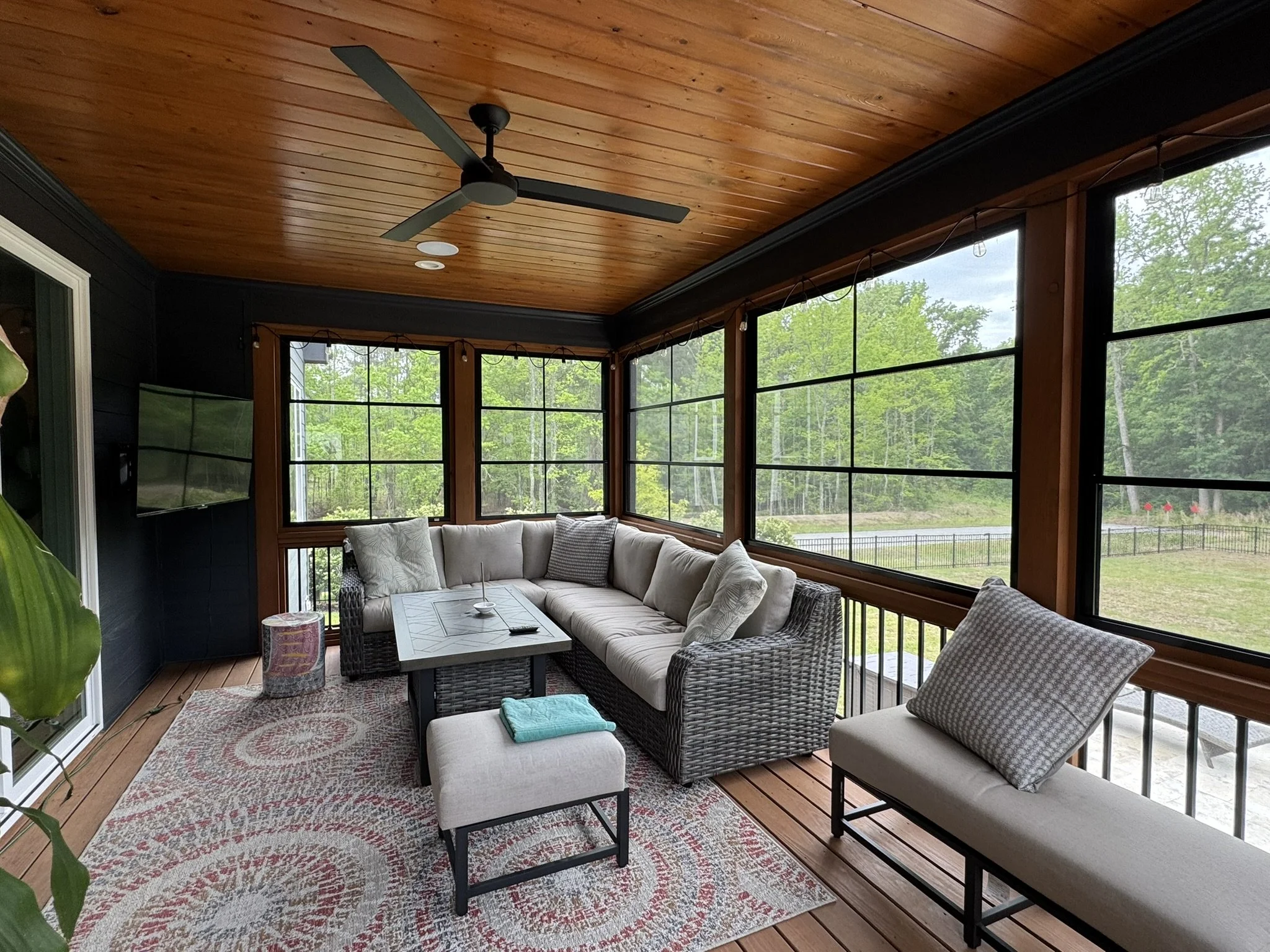 A screened porch with a wooden ceiling, ceiling fan, large windows showing a lush green landscape, and a cozy seating area with a white sectional sofa, pillows, a coffee table, and a patterned rug.