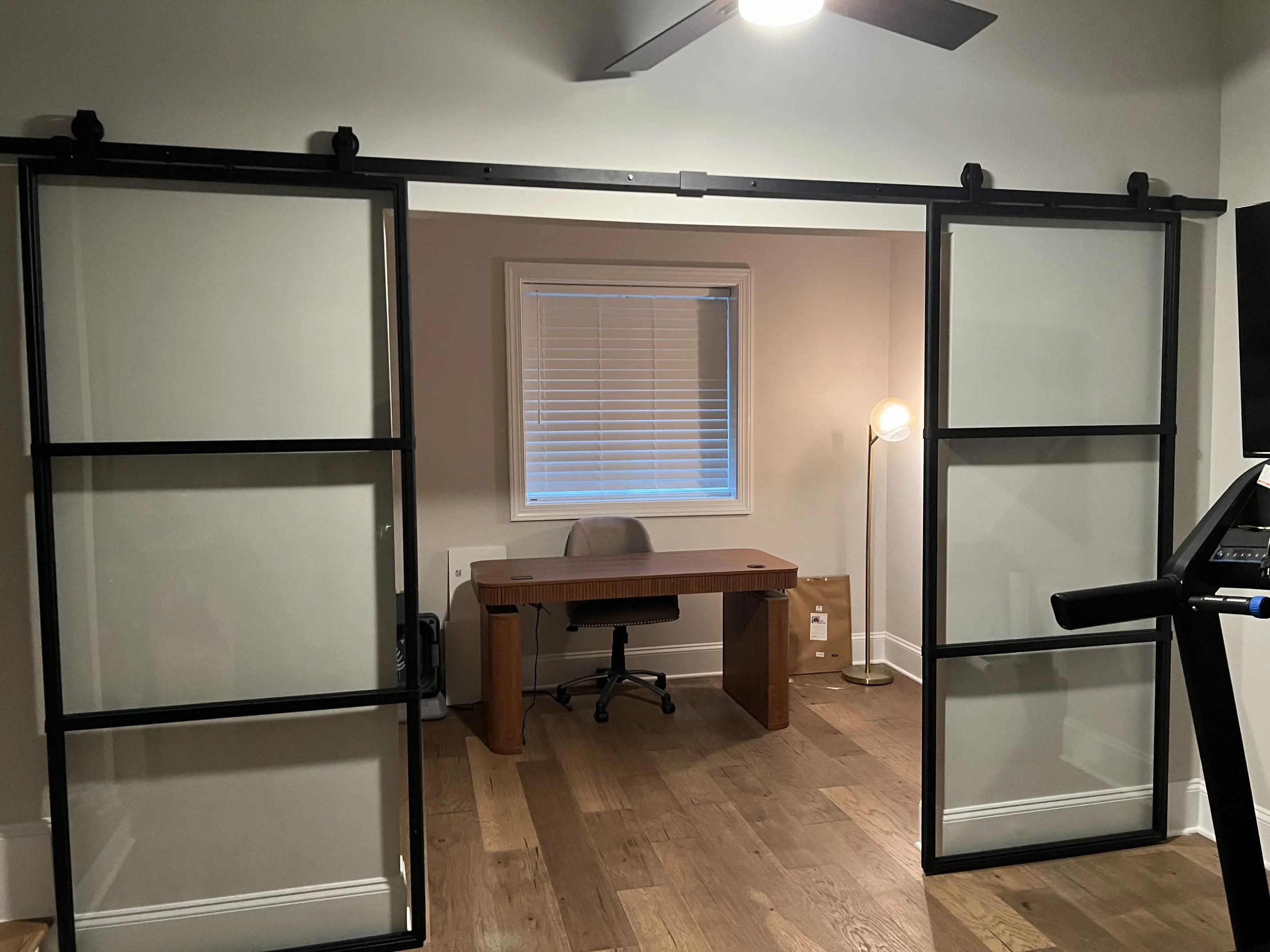 A room with two black metal and glass lofted bed frames in the foreground, a small wooden desk with a rolling chair, a window with closed blinds, a floor lamp, and part of a treadmill.