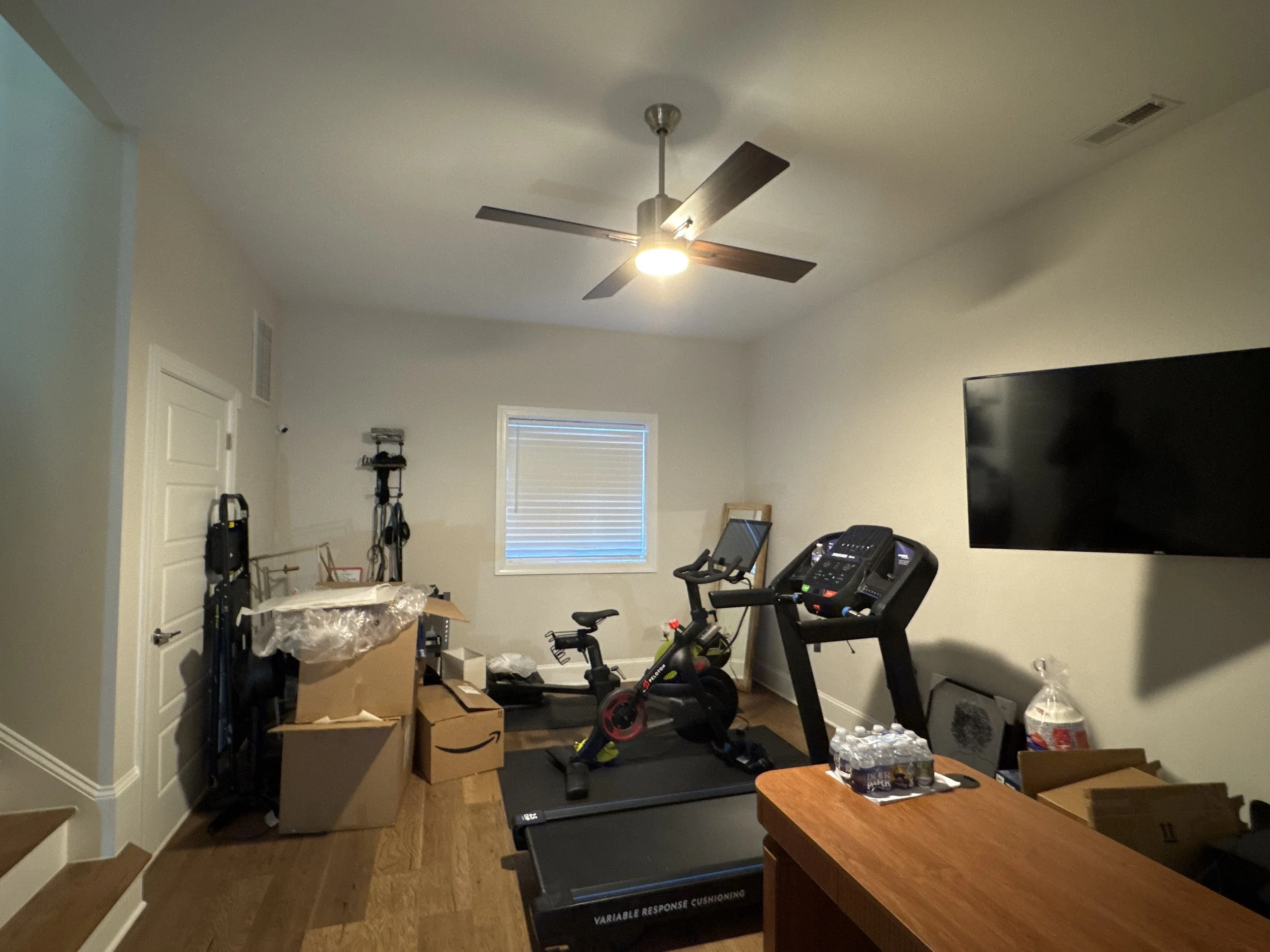 A room with a ceiling fan, workout equipment like a treadmill, and various boxes and items scattered around, including water bottles on a table.