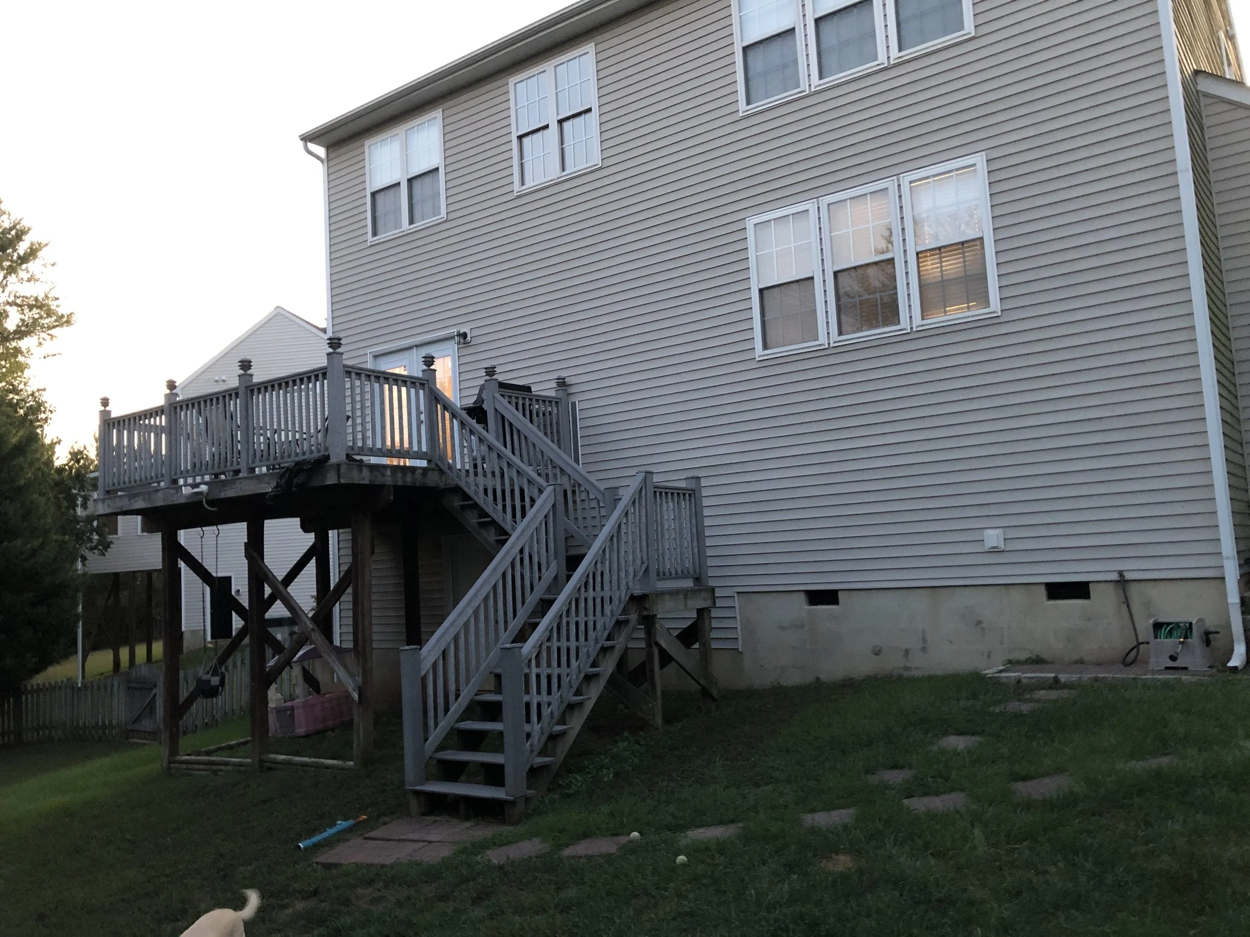 The image depicts the back of a beige multi-story house with several white-framed windows. There is a wooden staircase leading up to a small deck with a sliding glass door. The house has exterior siding and the yard has grass, a small section of ston