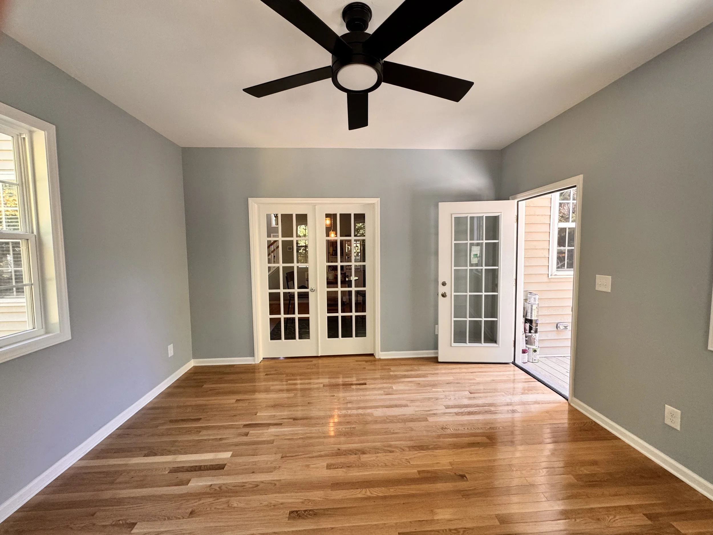 Empty room with hardwood floors, gray walls, white trim, and brown glass French doors leading to a dining area. There are windows on the left and right sides and a ceiling fan with black blades and a built-in light.