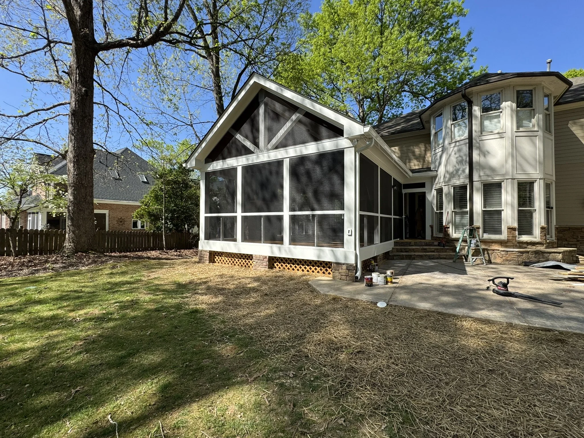 A backyard with a tree, a brick house, and a house with a stone foundation and tan siding. There is a screened-in porch being worked on with some tools and paint cans on the ground nearby. The sky is clear and blue.