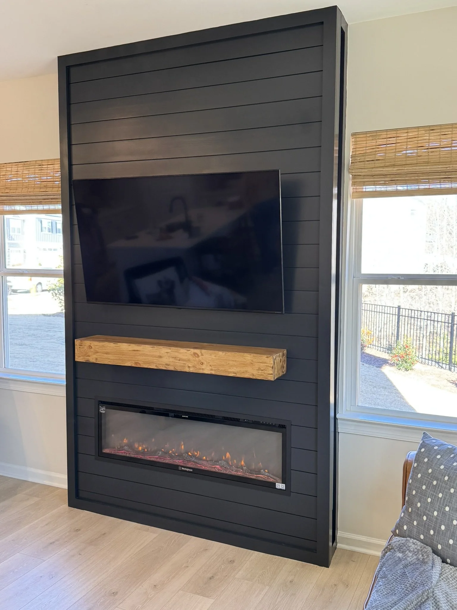 A flat-screen television mounted on a black slatted wall panel in a room with large windows, wooden floors, and a hanging electric fireplace. There is a partially visible polka-dotted chair with a gray blanket on it to the right.