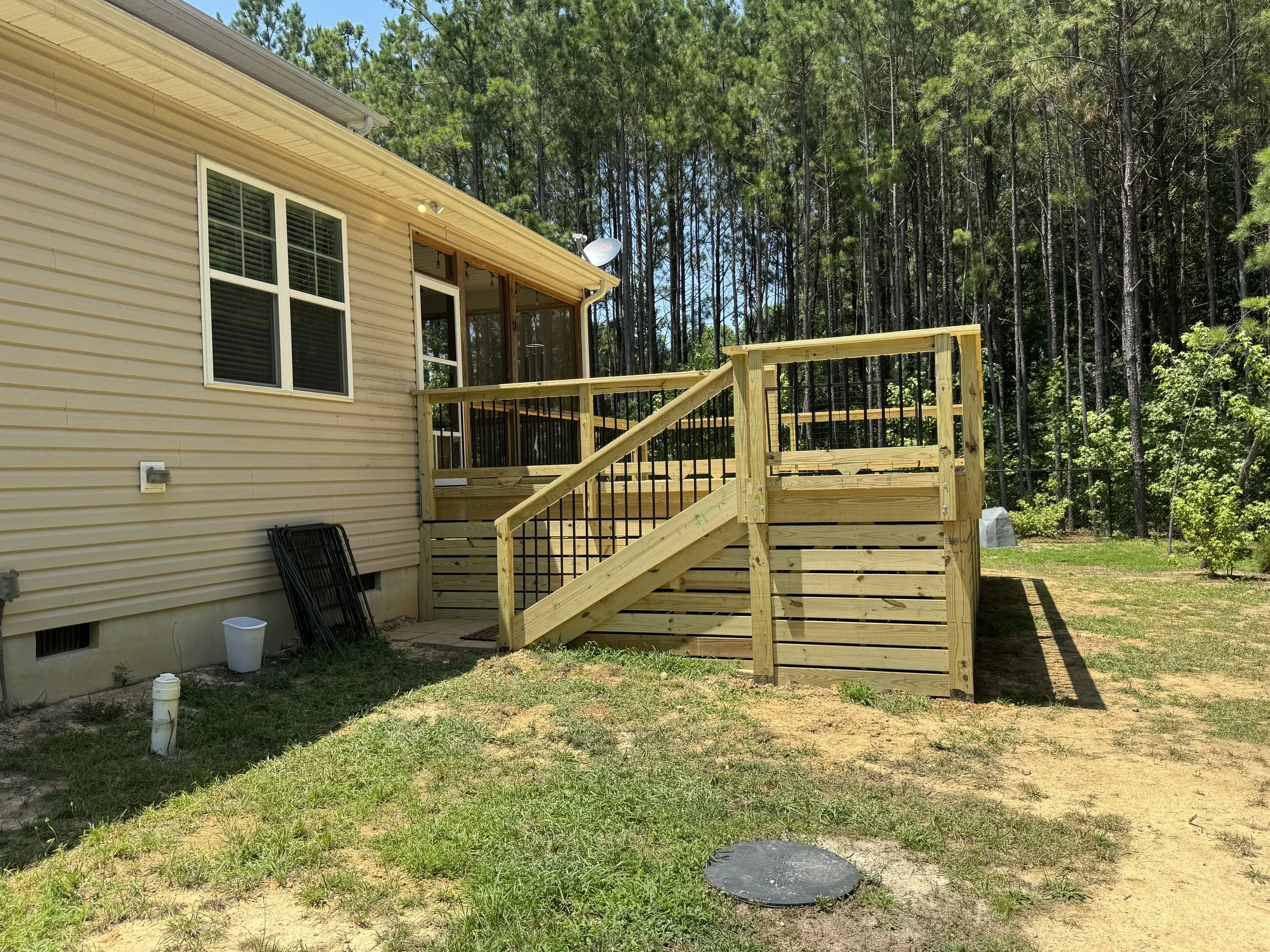 Backyard view of a house with beige siding, a new wooden deck with stairs, and a wooded backyard with tall pine trees.
