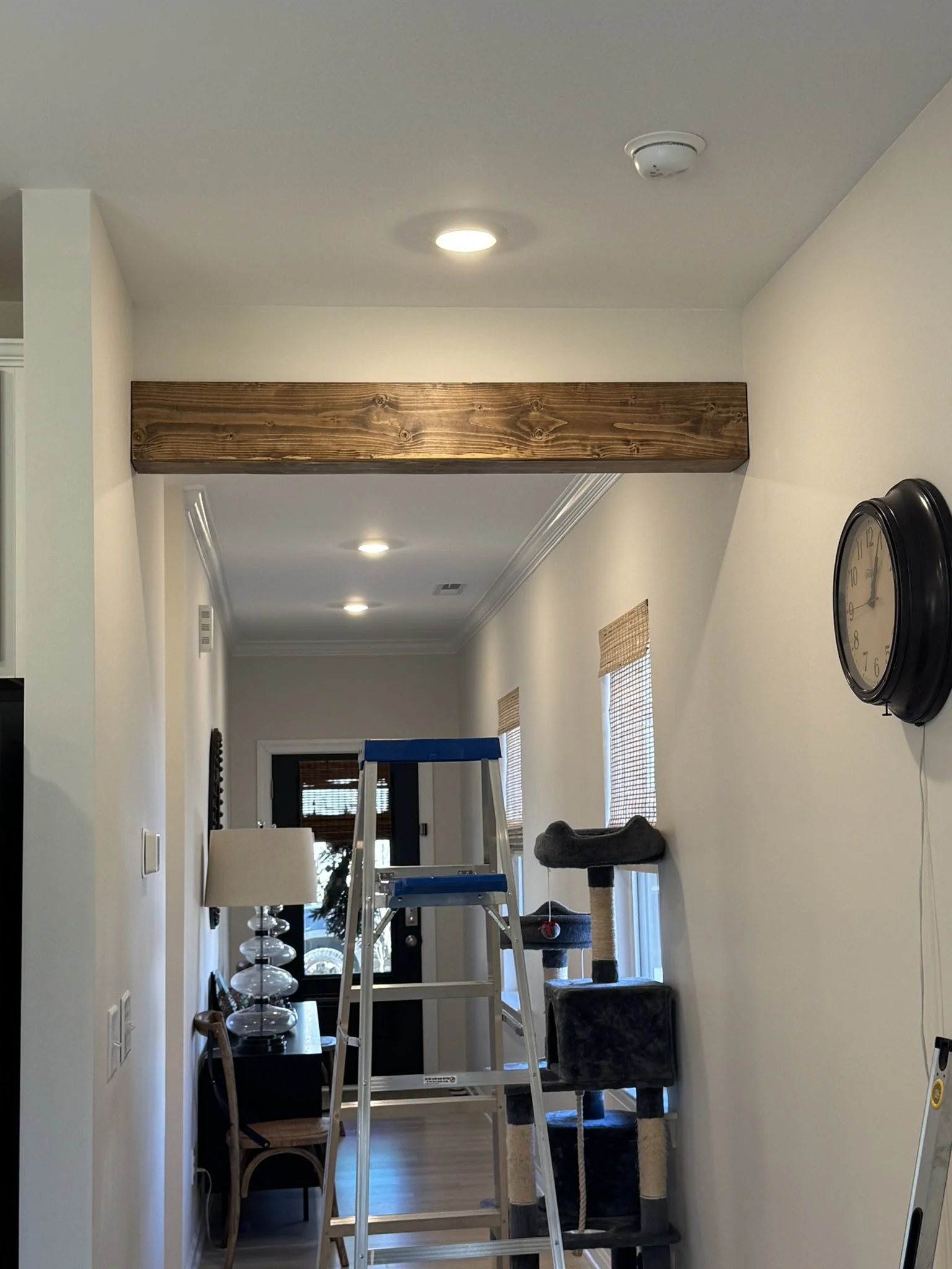 A hallway inside a house under renovation, with a wooden beam across the ceiling, a stepladder, and various furniture and decorations, including a dark-colored cat tree, a lamp, and a wall clock.
