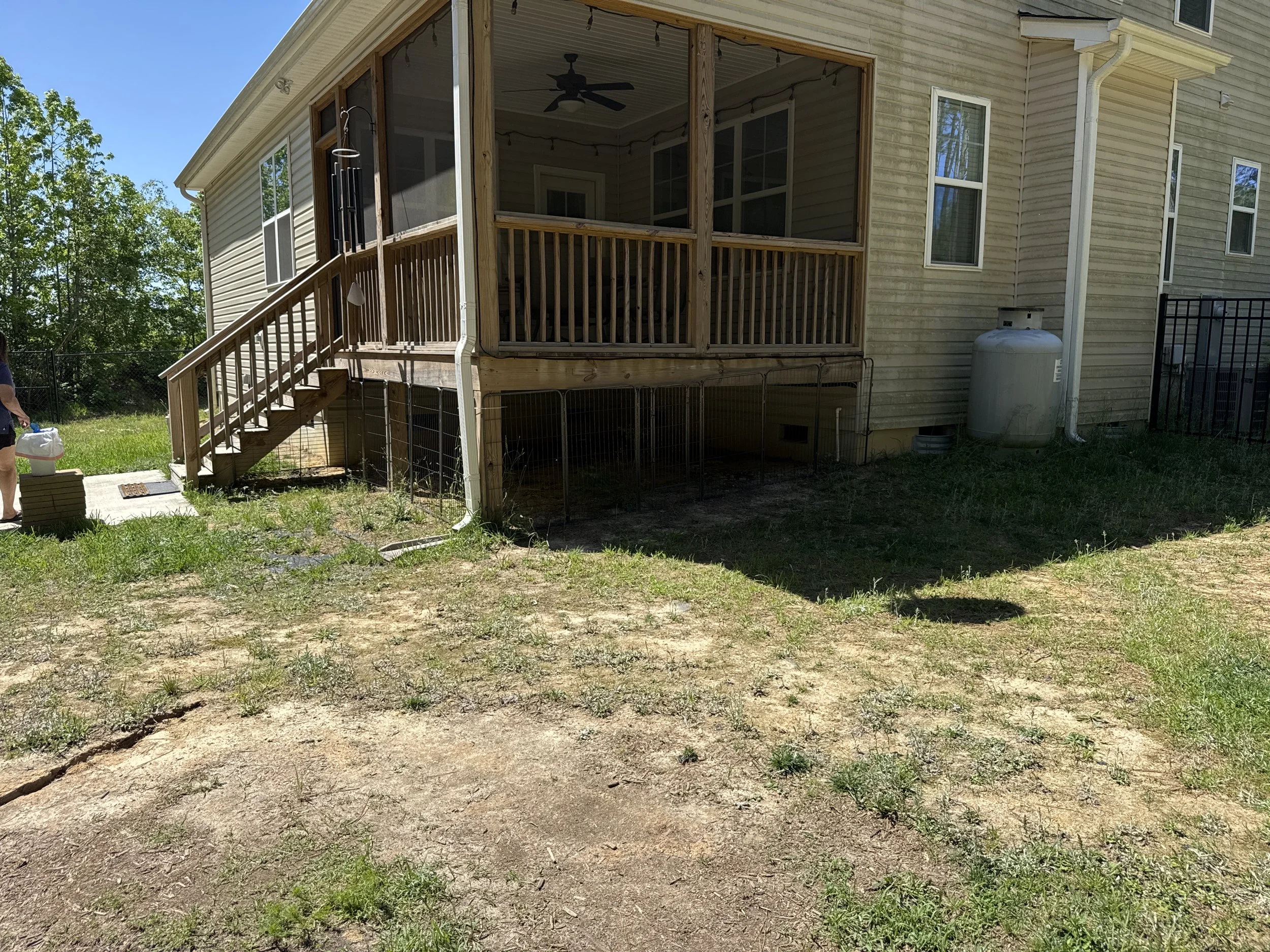 A backyard with a newly built screened-in porch attached to a house, with a wooden staircase leading up to it. There is a propane tank to the right of the porch and a small man-made grassy area with some patches of dirt in the foreground. The sky is 