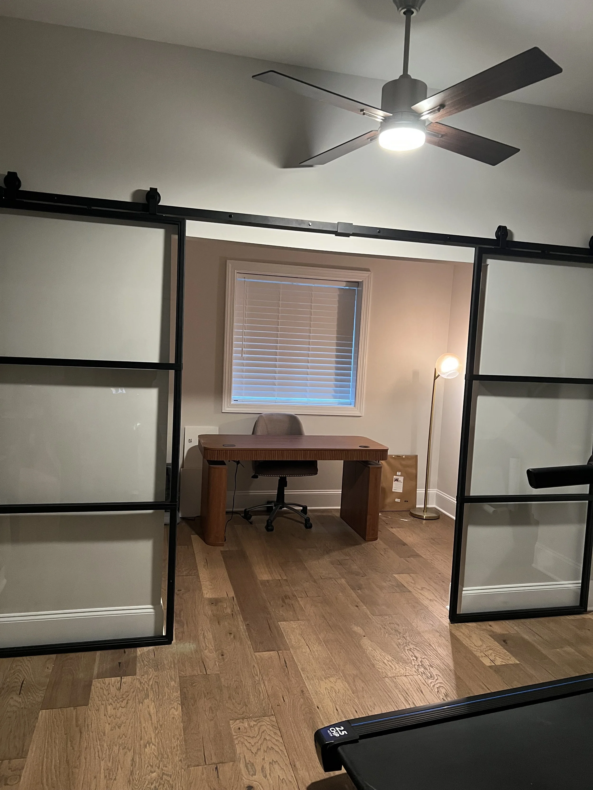 A home office room featuring a wooden desk with a gray office chair, a window with blinds, a floor lamp with a spherical shade, a ceiling fan with wooden blades and a bright light, and sliding glass doors with frosted glass panels.