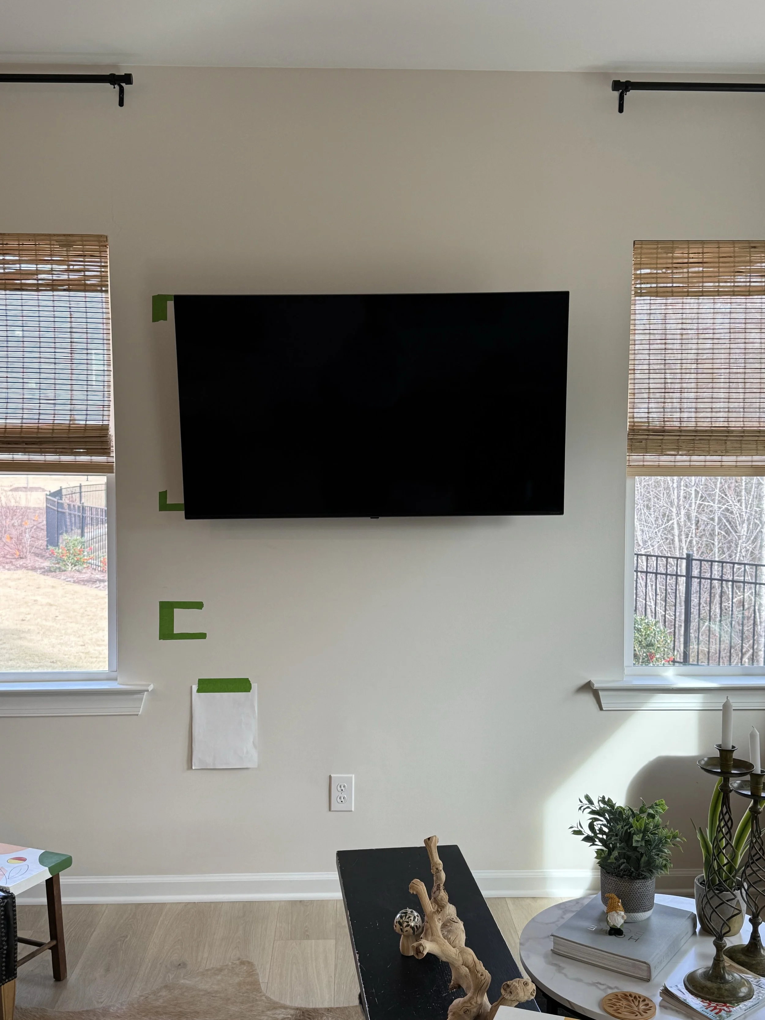 Living room wall with a large flat-screen TV mounted above a small table, two windows with bamboo blinds, and decorative items on a round table including potted plants, books, and candles.