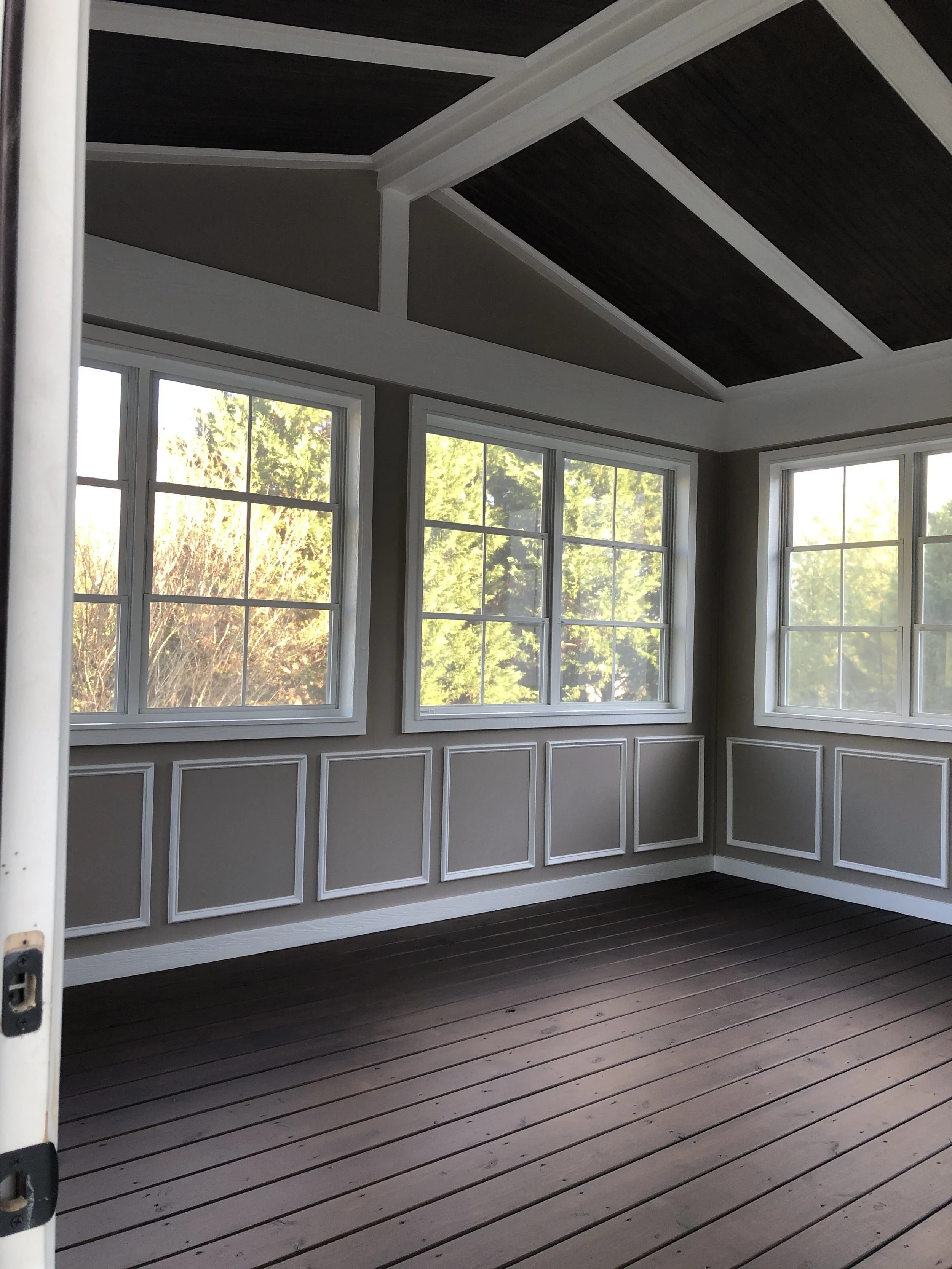 Empty room with large windows, gray walls with white wainscoting, and dark wood ceiling panels.