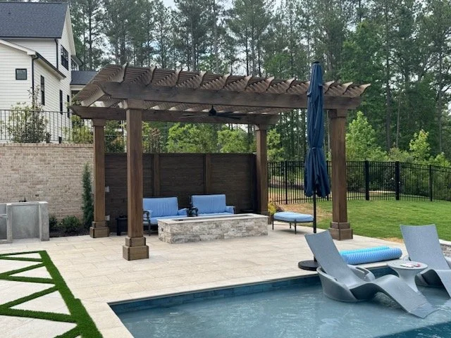 Backyard patio area with a pergola, outdoor seating, a fire pit, lounge chairs by a swimming pool, and a grassy yard enclosed by a black fence.