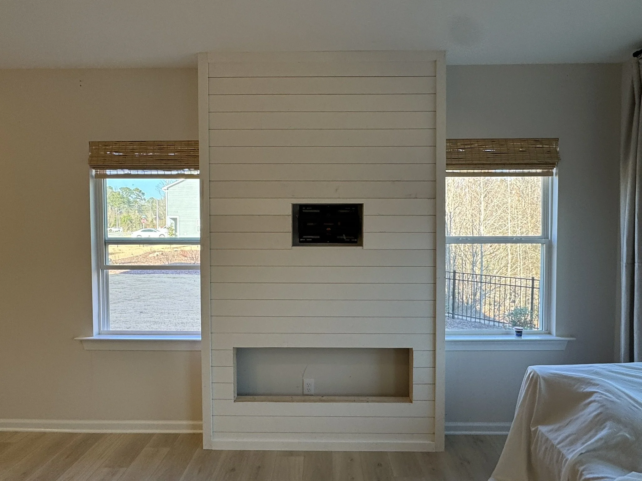 Empty room with white shiplap wall, two windows with bamboo shades, and a small fireplace opening in the wall. Bed partially visible on the right, with a small container on the window sill.