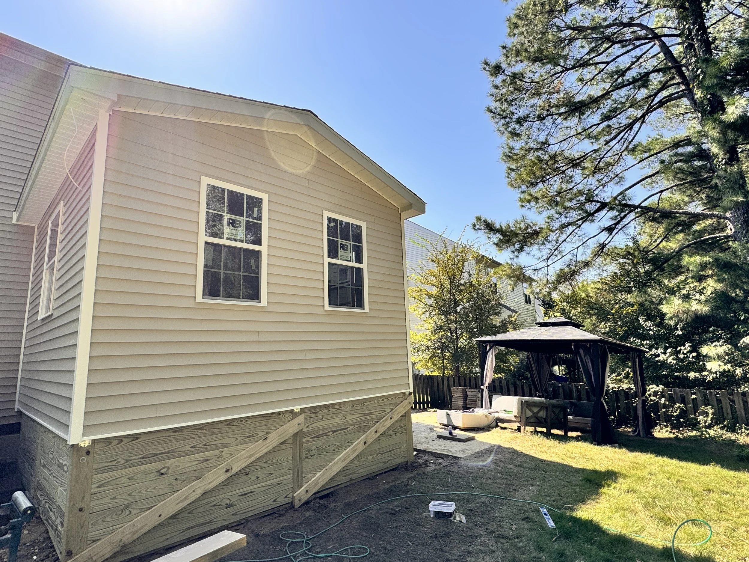 A house with beige siding and white trim, under construction, with a raised wooden deck and a backyard with a black gazebo and trees.