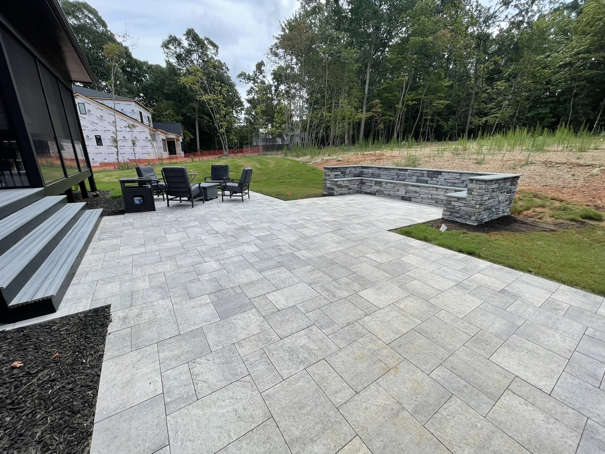 Backyard with patio furniture, stone retaining wall, grass lawn, and trees in the background, with an under-construction house beside a wooded area.