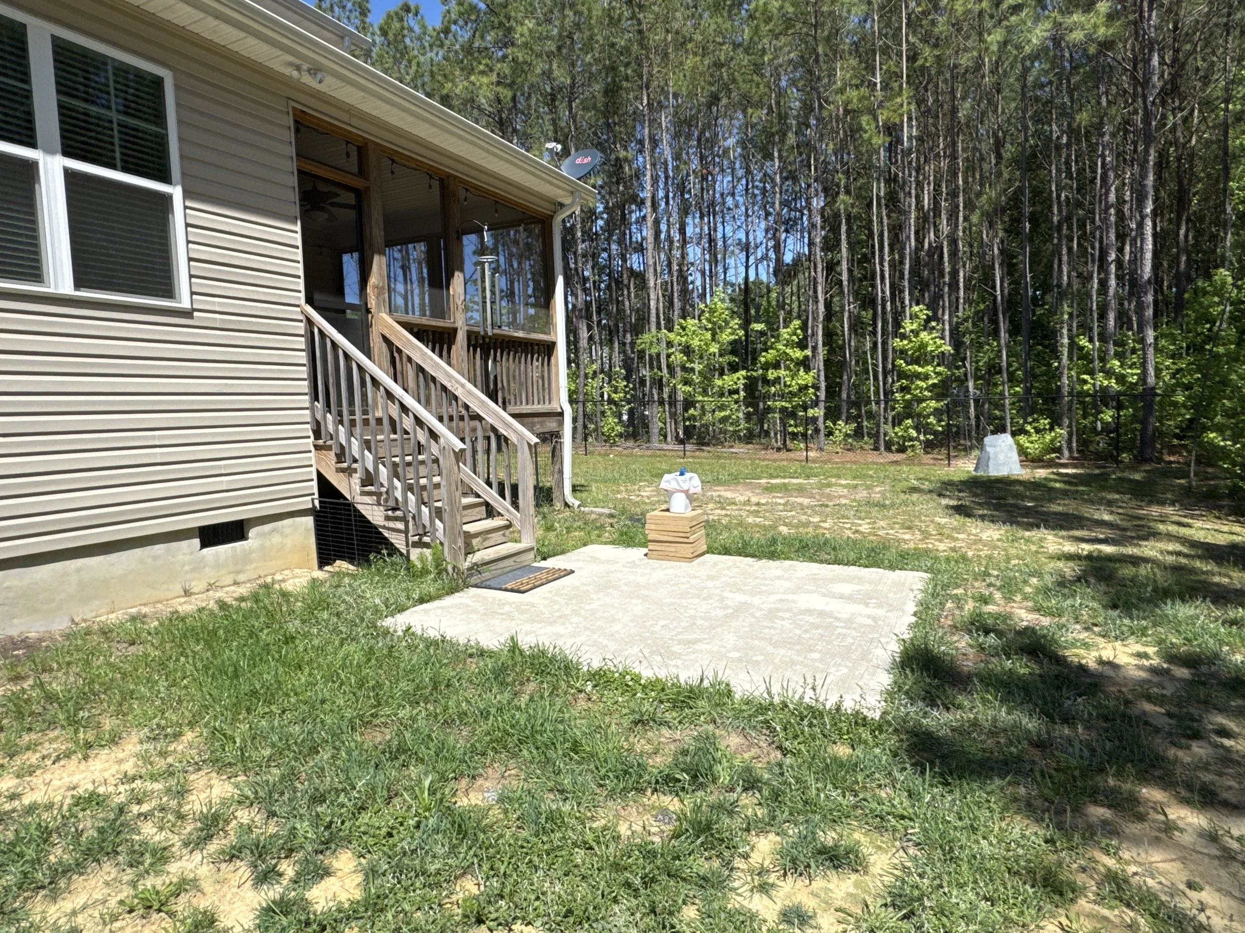 Exterior view of a house with beige siding, a screened porch, and a staircase leading to the yard. There are trees and a fence in the background, along with a satellite dish on the roof. The yard has grass, a bird feeder, and a stone decorative piece