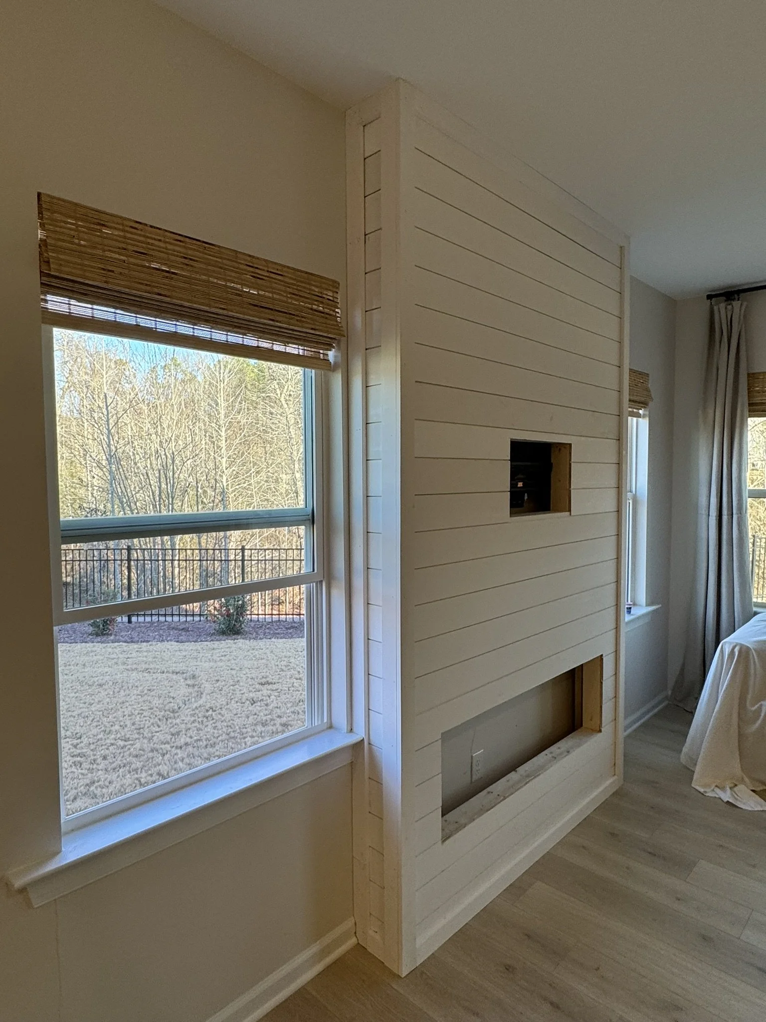 Interior of a room under renovation with a partially built wooden fireplace and two windows with bamboo blinds, showing an outdoor yard and trees.
