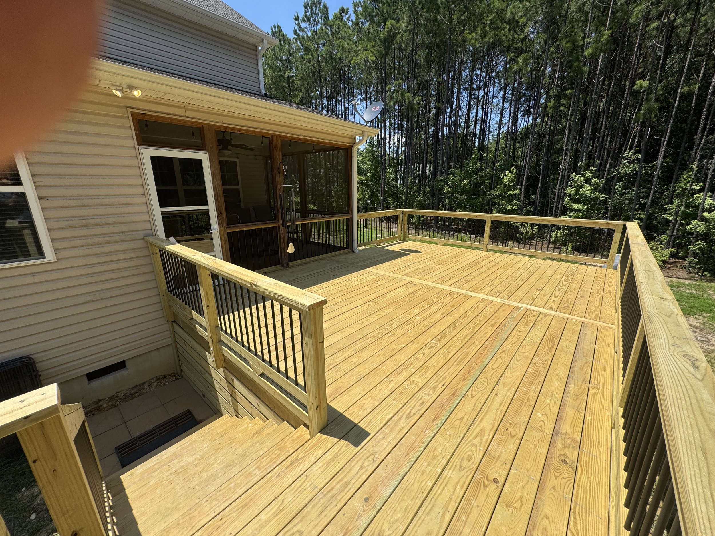 Newly built wooden deck attached to the back of a house, with black metal railing and a screened porch area, surrounded by a forest of tall trees.