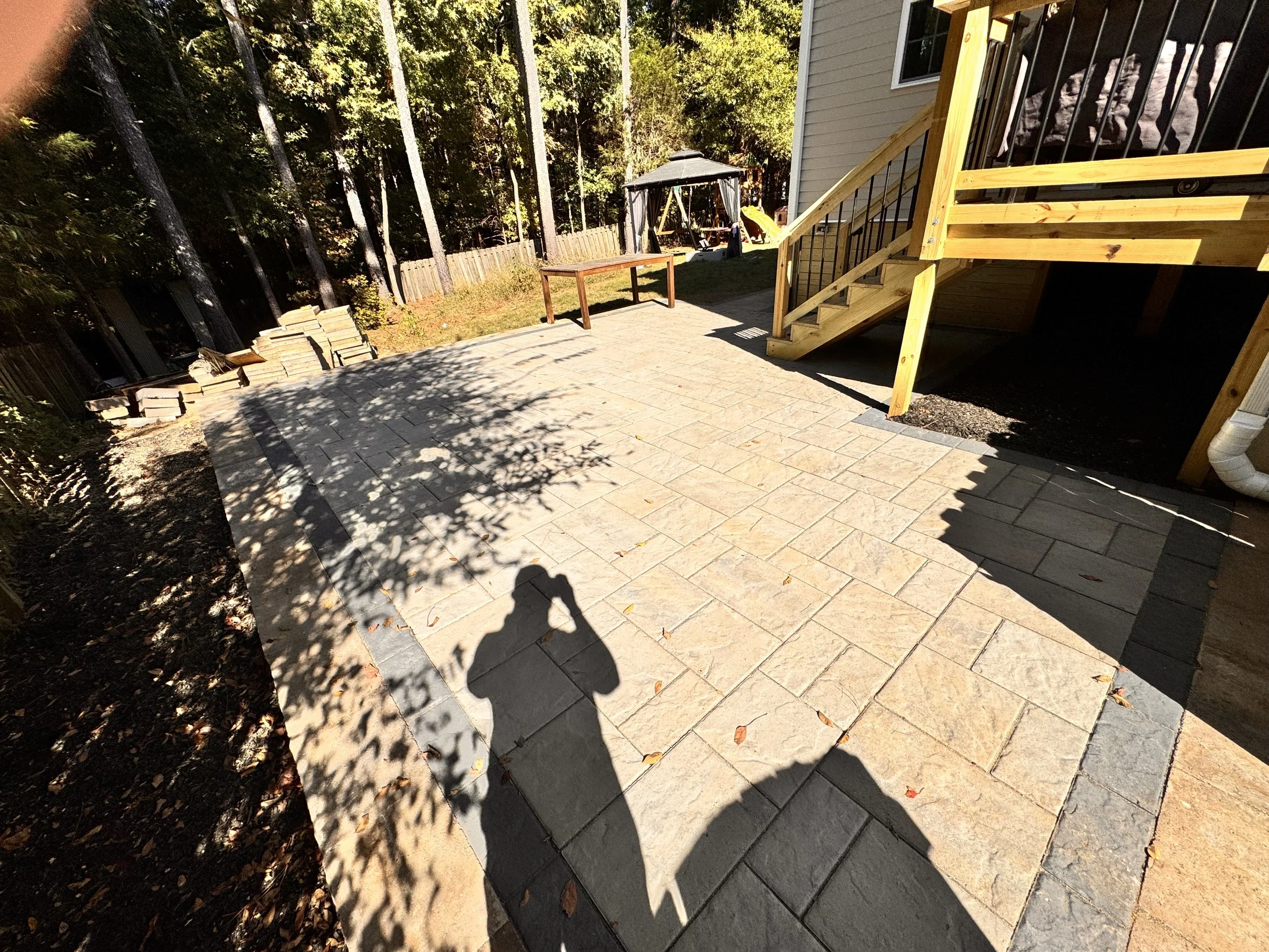 Backyard patio area with stone pavers, a wooden staircase leading to an upper deck, a table, and a swing set in a wooded area with trees and a fence.