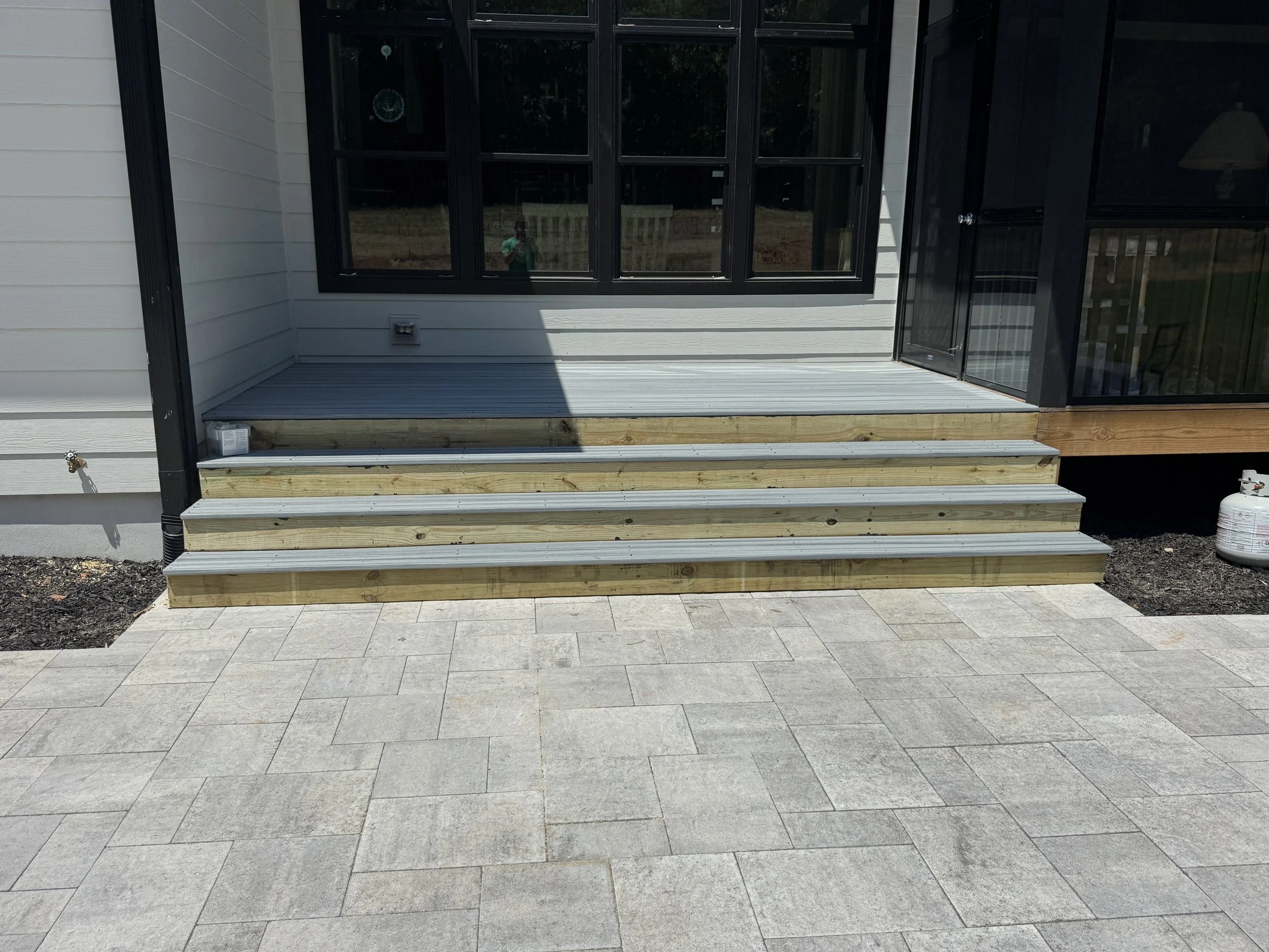 A house porch with steps leading up to a screened-in porch area. The porch has black framing around the screen and beige side walls. There is a small sink or water connection on the left side near the ground and a propane tank on the right side, all 