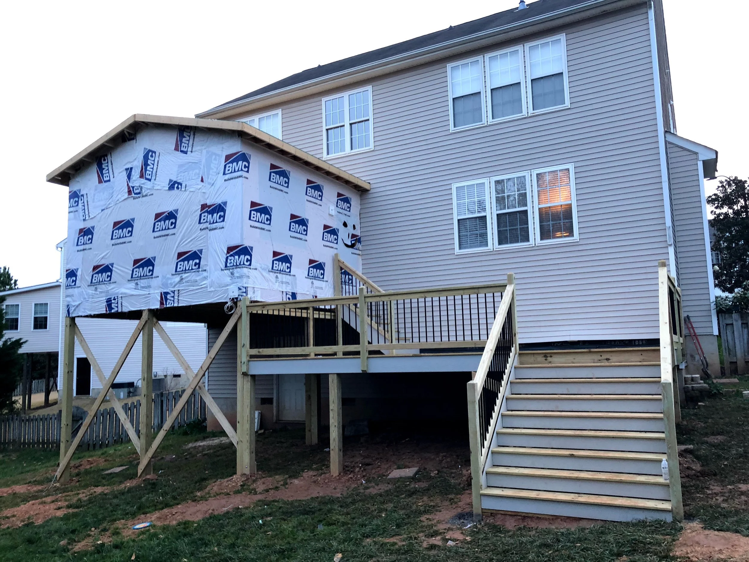 A house under construction with beige vinyl siding, multiple windows, and a wooden staircase leading to a second-story deck. There is also a small attached structure wrapped in BMC building wrap.