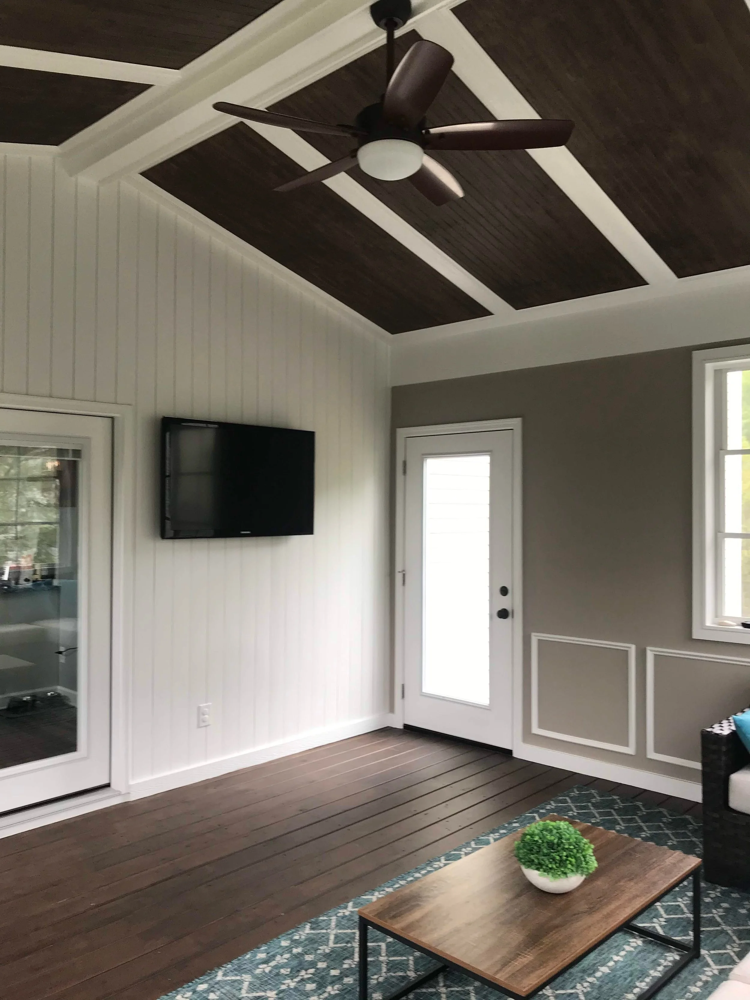 Living room with a ceiling fan, a wall-mounted television, a glass door, windows, a wooden coffee table with a decorative bowl of green plants, a patterned rug, and a partial view of a grey wall with white paneling.