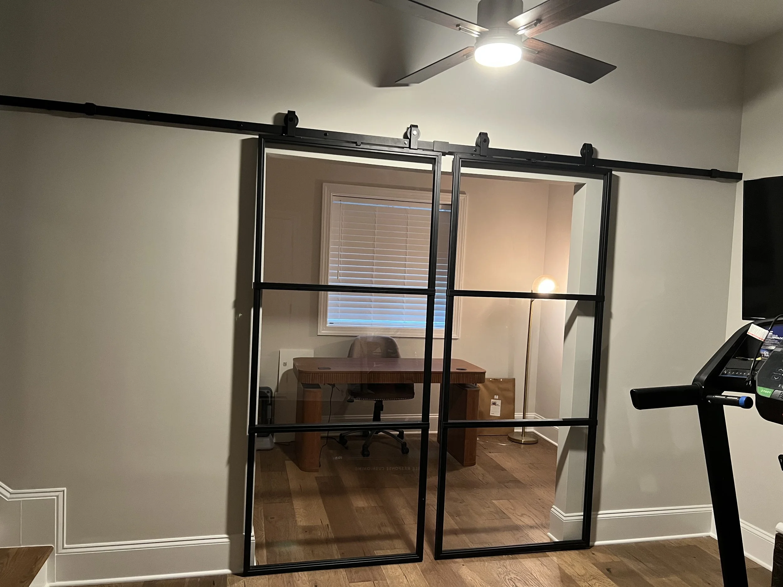 A room with a ceiling fan, a window with blinds, a desk, a chair, a floor lamp, and a treadmill.