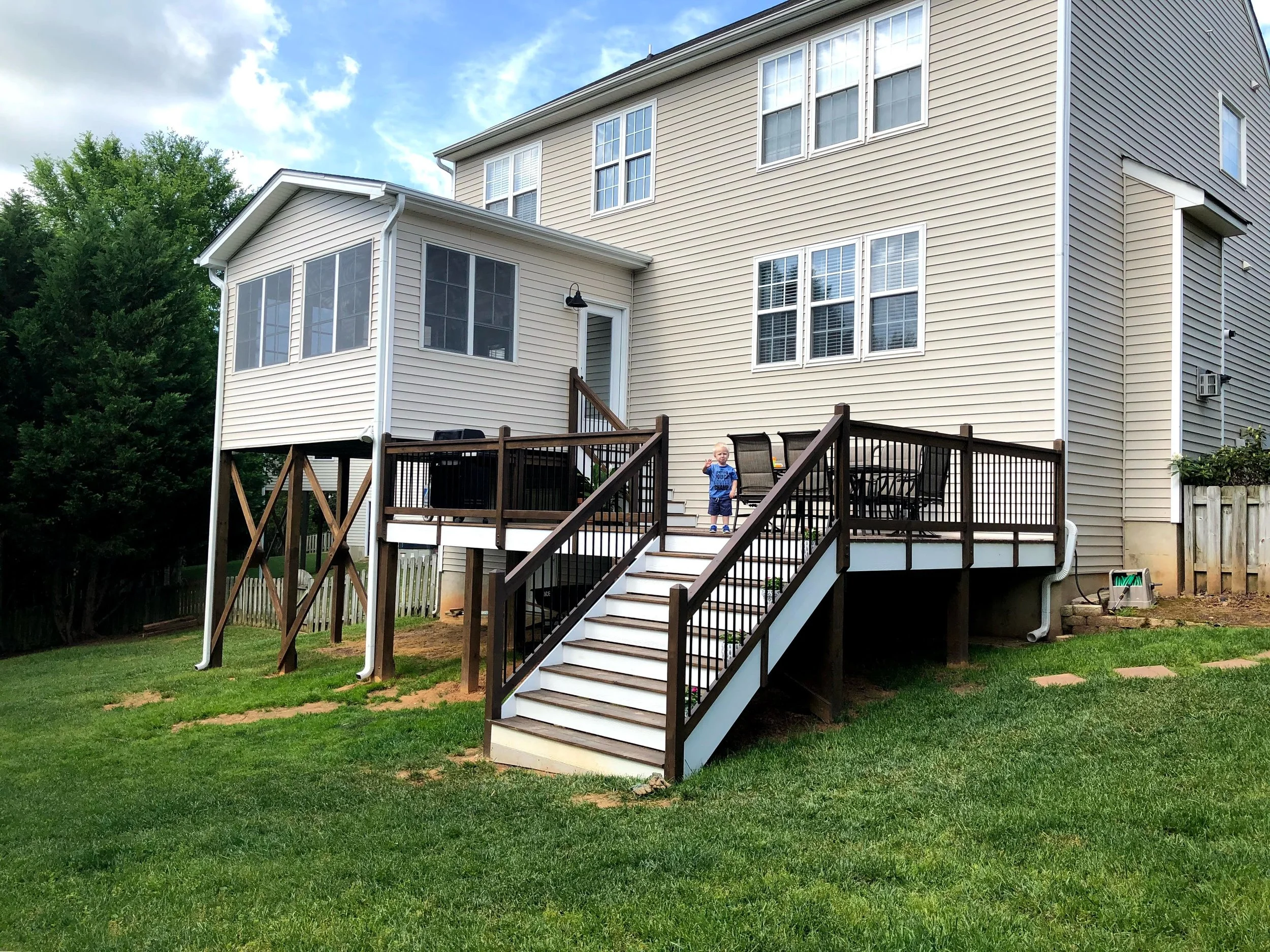 A backyard view of a beige multi-story house with a wooden deck and stairs, a child standing on the deck, green grass, trees, and a partly cloudy sky.