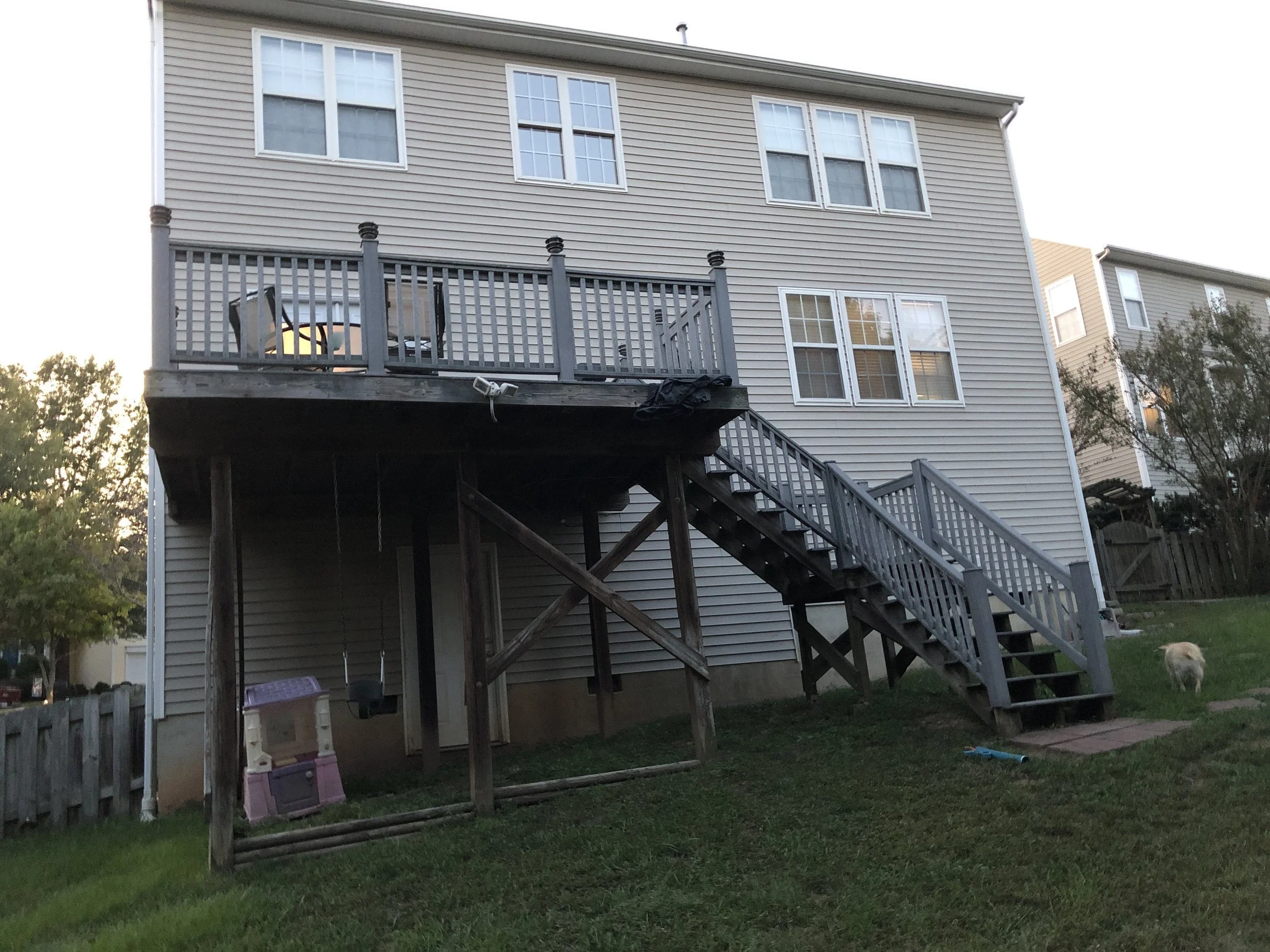 Backyard of a house with a deck and stairs leading down to the grass, and a dog near the bottom. The house has beige siding and multiple windows, with a swing hanging from the deck.