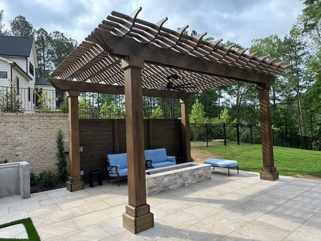 Outdoor patio area with a wooden pergola, built-in seating with blue cushions, and a square fire pit centerpiece on a tiled floor, with trees and a fence in the background.