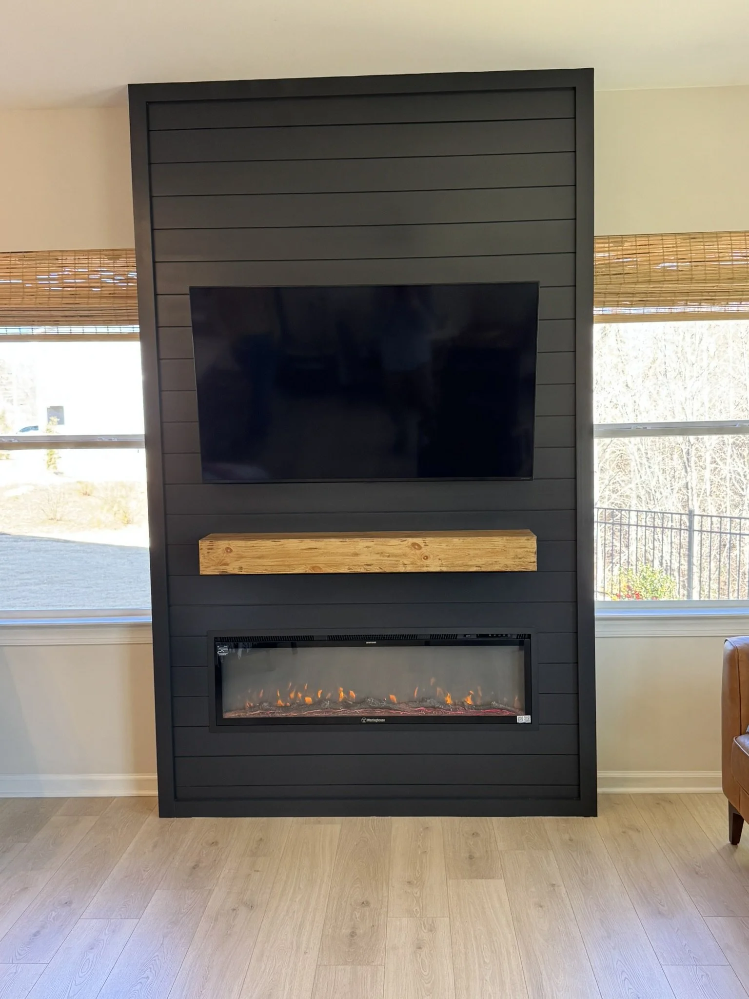 Living room with a black TV over a modern electric fireplace, flanked by two windows with bamboo blinds, and light hardwood flooring.