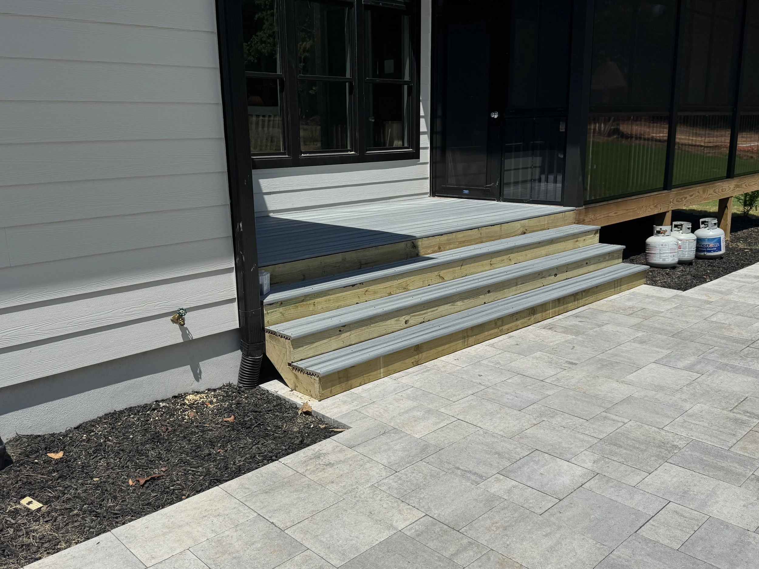 Newly built house porch with stairs, gray siding, black trim, black screened door, and propane tanks nearby.