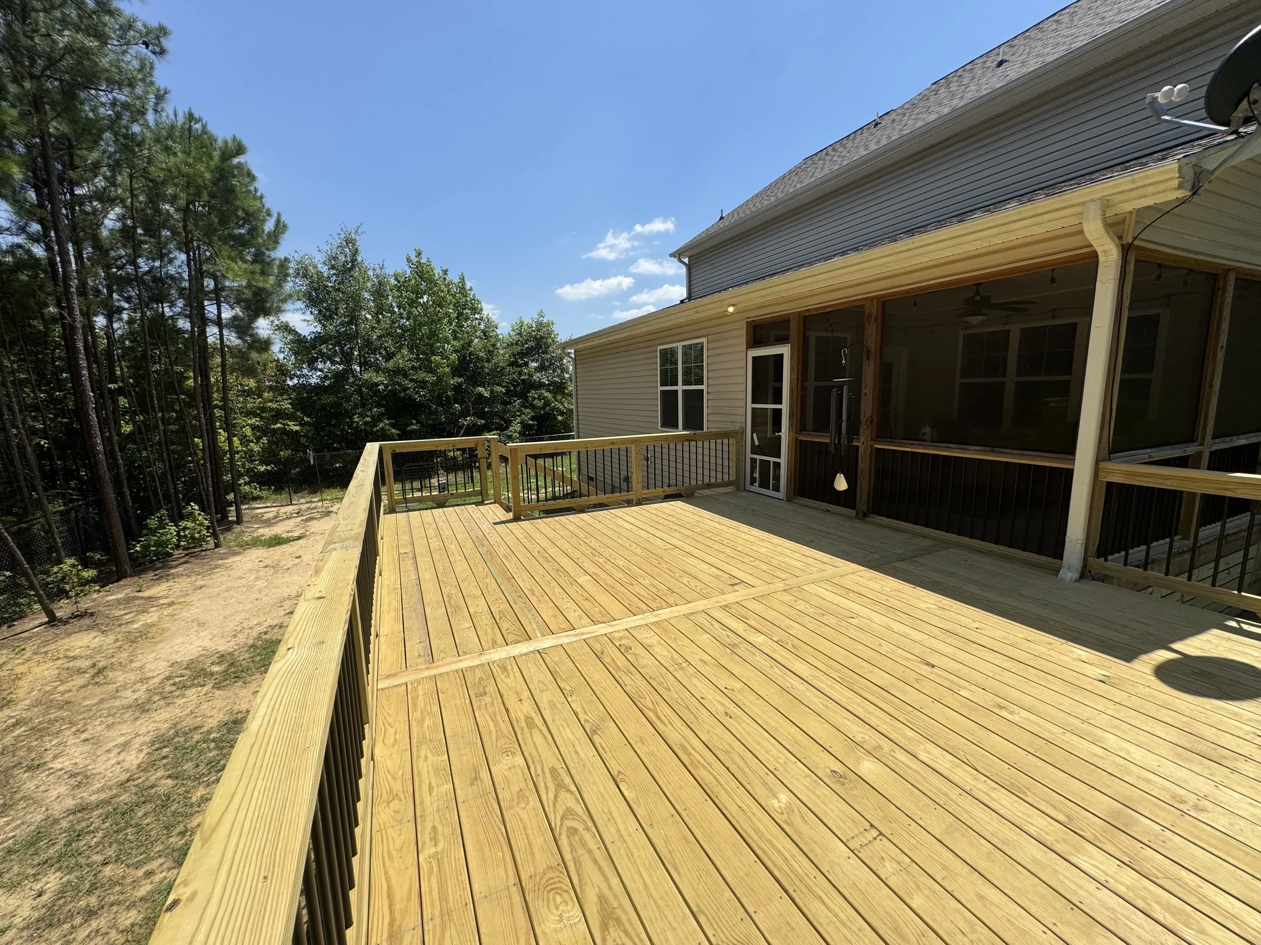 Newly built wooden deck attached to a house, with a safety railing, overlooking trees and a blue sky with a few clouds.