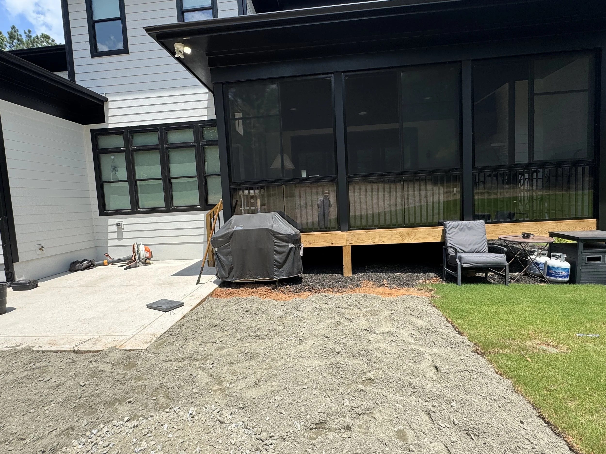 An outdoor patio area adjacent to a house with black-framed windows, a screened porch, and some outdoor furniture. The ground is partially covered with gravel and partially with grass.