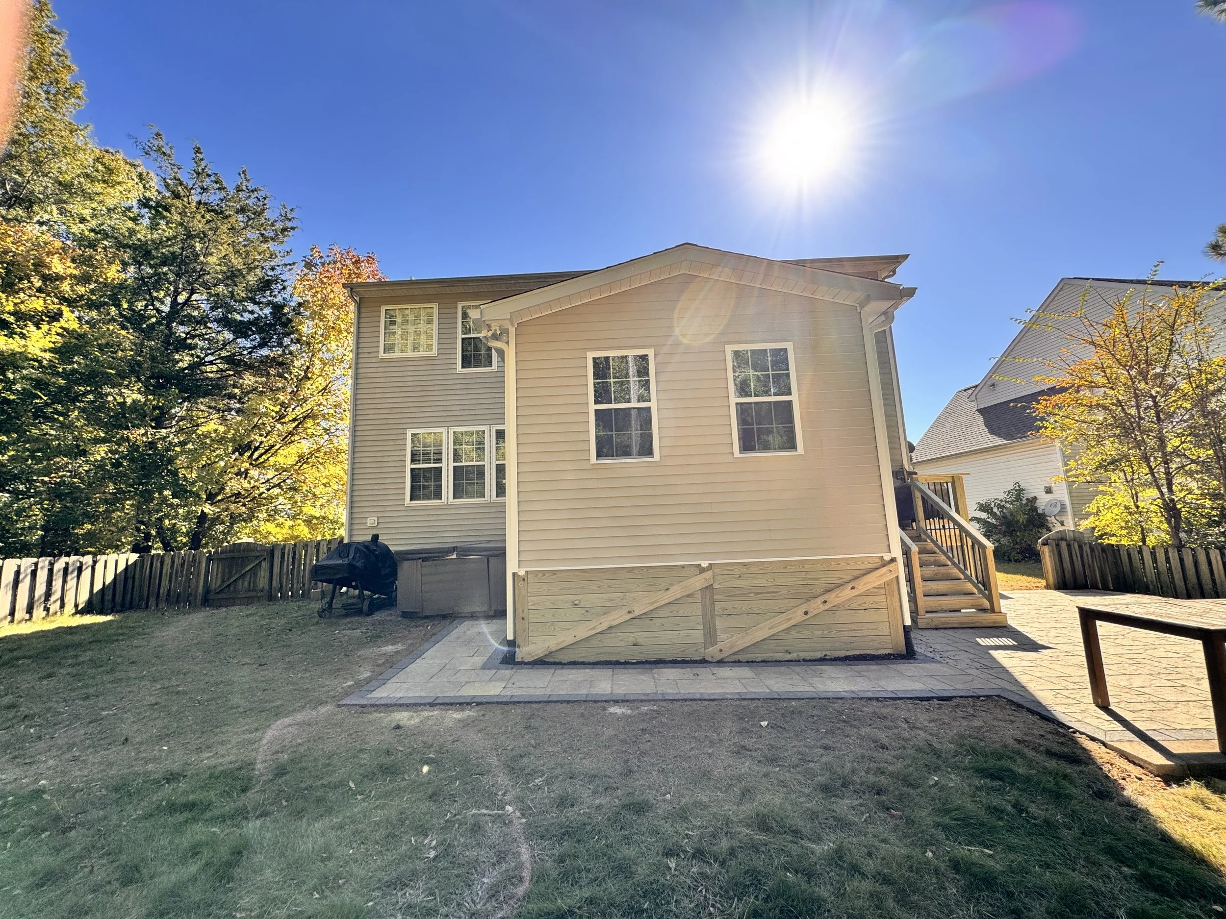 A backyard with a house, trees, a paved patio, a grill, and a wooden staircase leading to a deck. The sky is clear with the sun shining brightly.