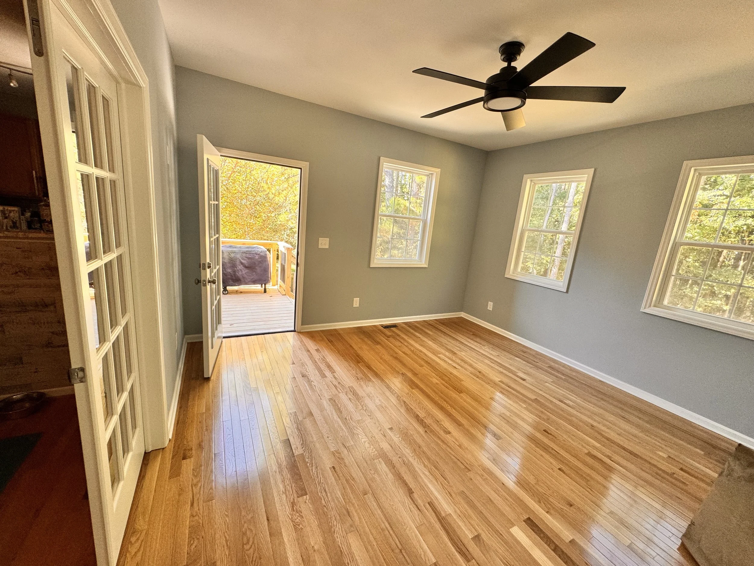 Living room with hardwood floors, light gray walls, multiple windows letting in natural light, a ceiling fan, and a door leading to a wooden deck outdoors.