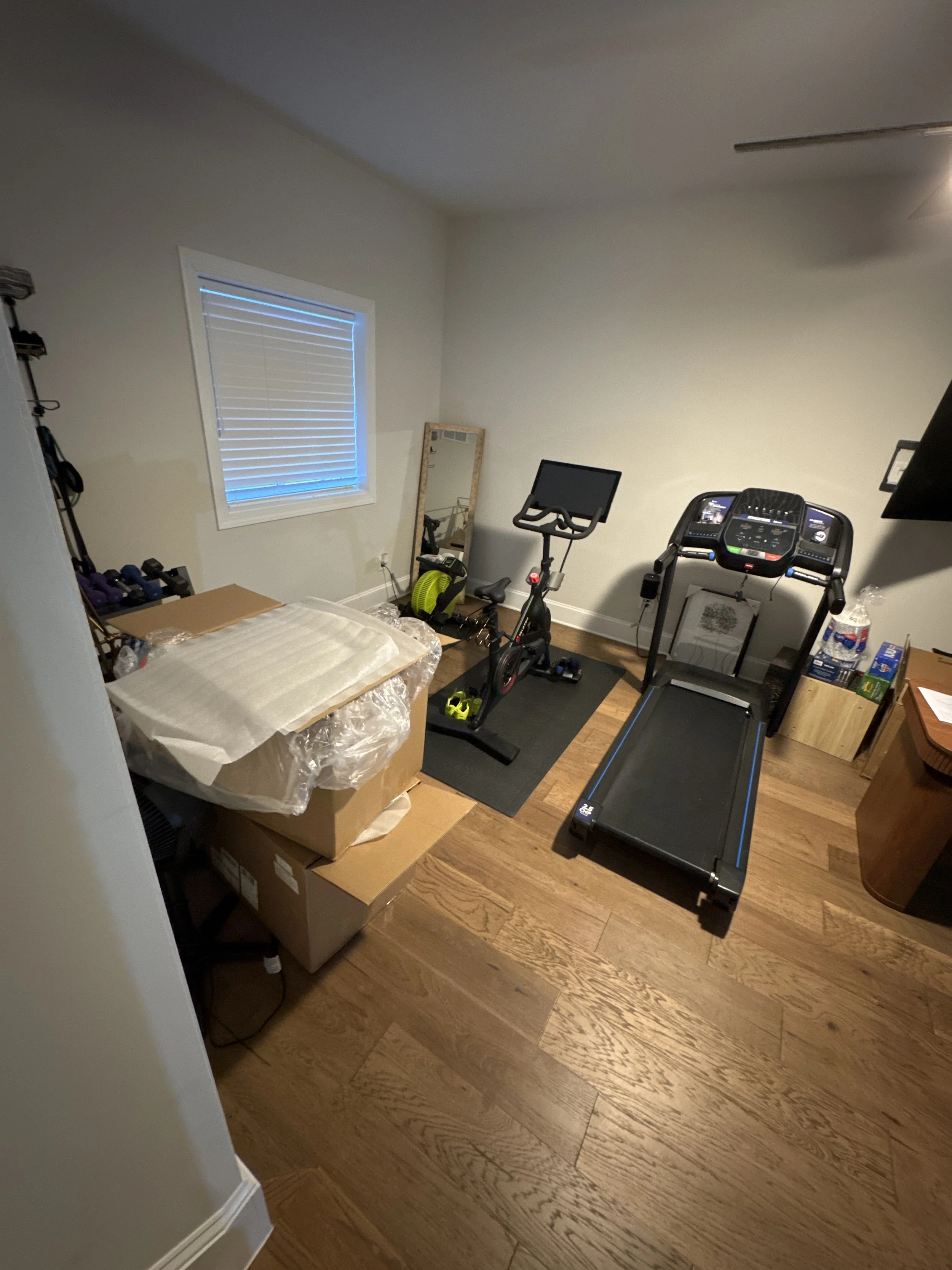 A room with a wooden floor, white walls, and a ceiling fan. There is a treadmill on the right side, a stationary bike in the center, a mirror leaning against the wall, some boxes and gym equipment, and a window with closed blinds.