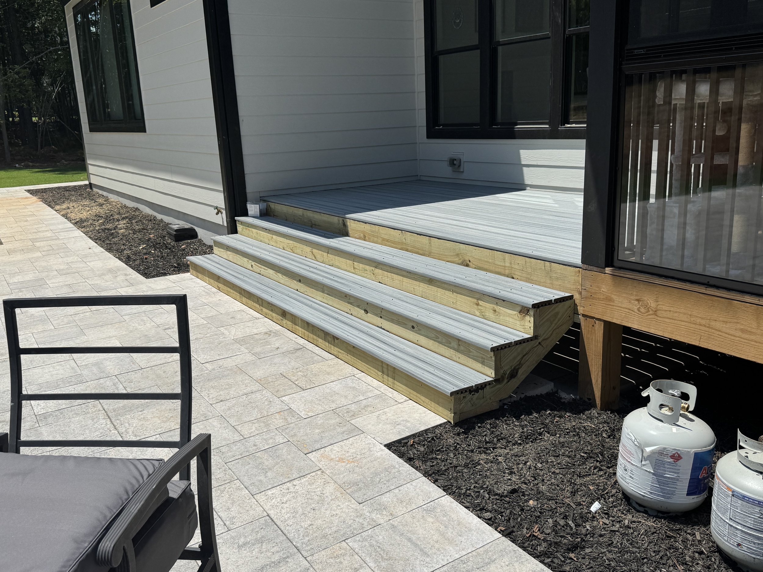 A partially constructed wooden deck attached to the back of a house with gray siding and black-framed windows. Two steps lead up to the deck, which is in the process of being built, with some tools and propane tanks nearby.