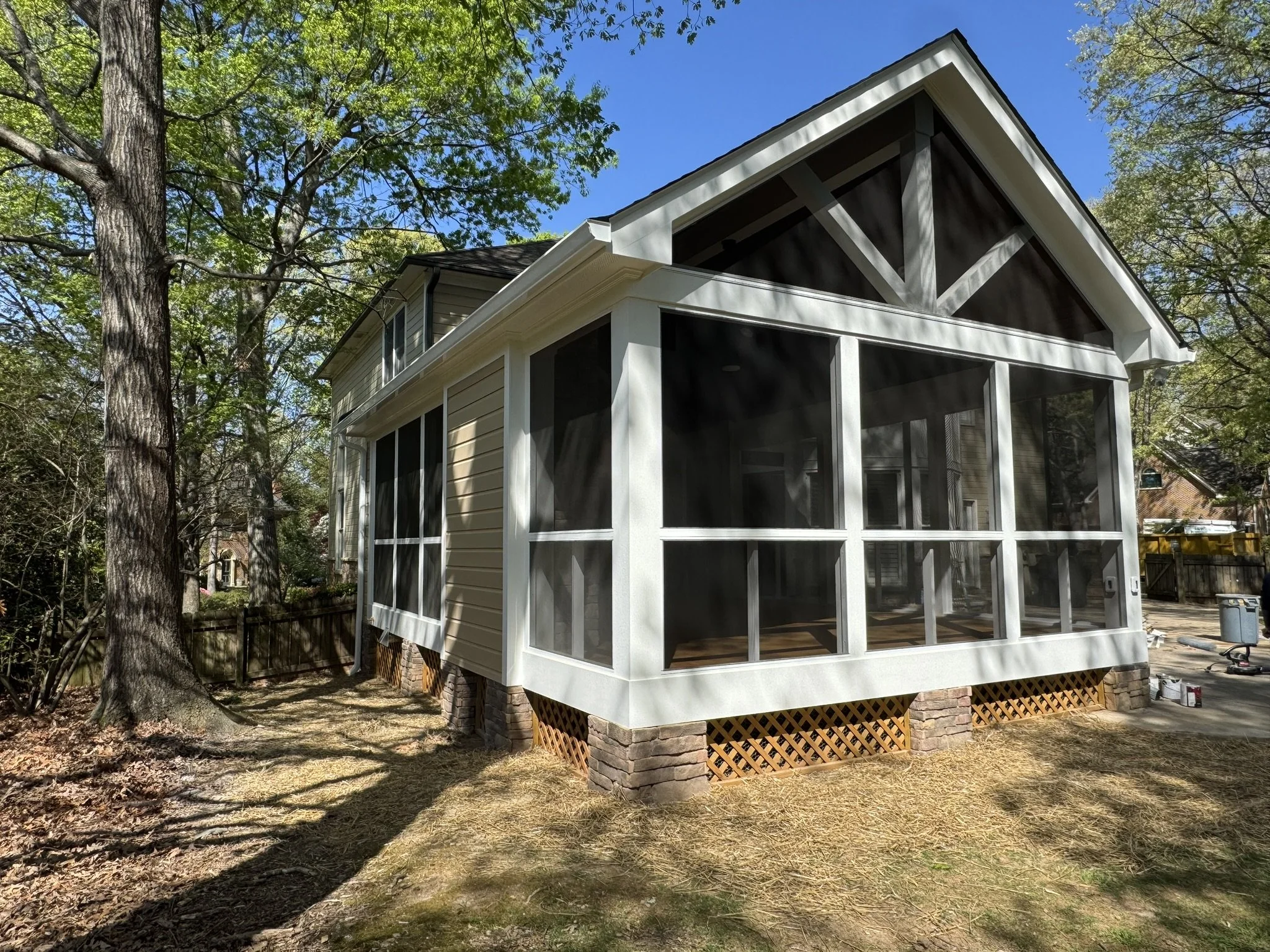 A house with a screened porch is elevated on brick pillars in a backyard surrounded by trees and a dirt yard.