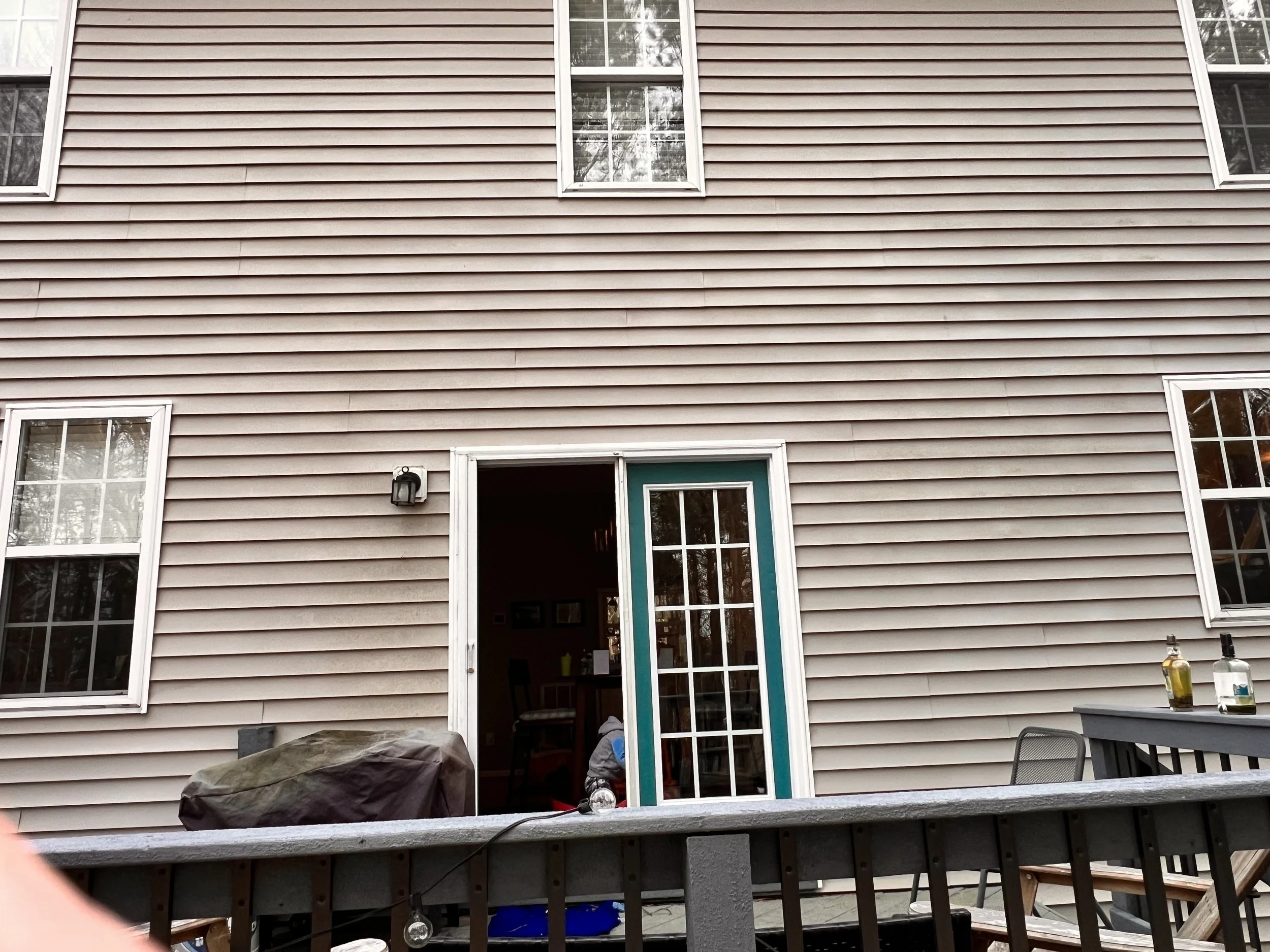 Backyard with house siding, four windows, and sliding glass door with open door. Patio furniture, barbecue grill covered, and some bottles on table.