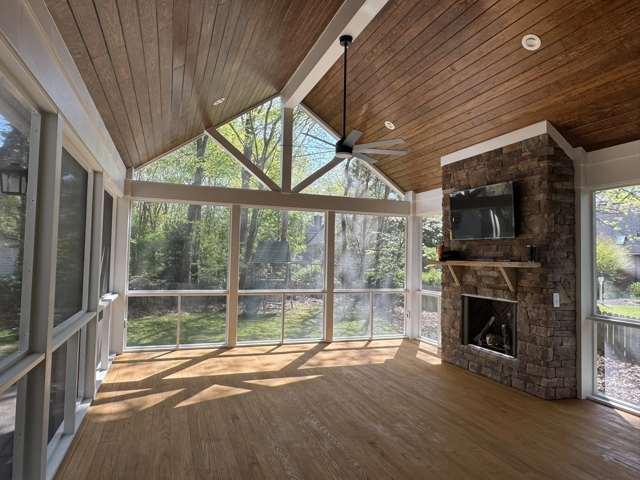 Living room with a stone fireplace, large windows, and a ceiling fan with multiple blades. Sunlight and trees are visible through the windows.