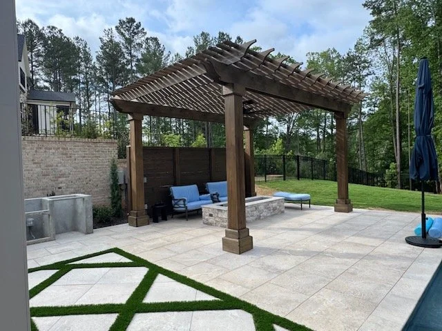 Outdoor patio area with a wooden pergola, blue seating, a fire pit, an umbrella, and a grassy yard with trees in the background.