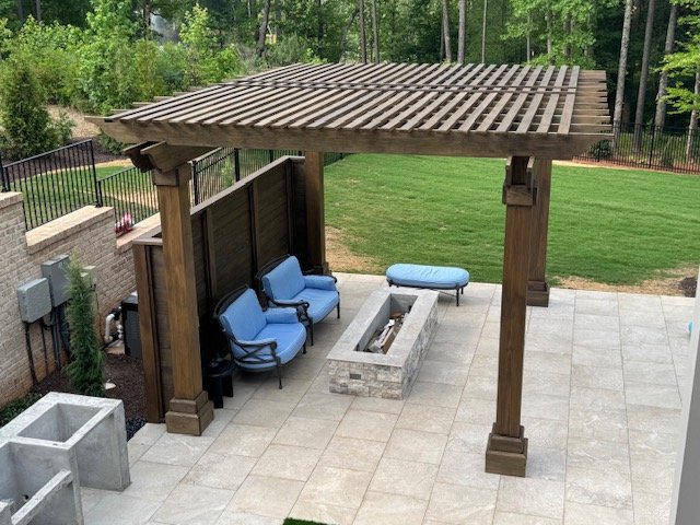 Backyard patio with a wooden pergola, blue outdoor chairs, and a fire pit, surrounded by green grass and trees.