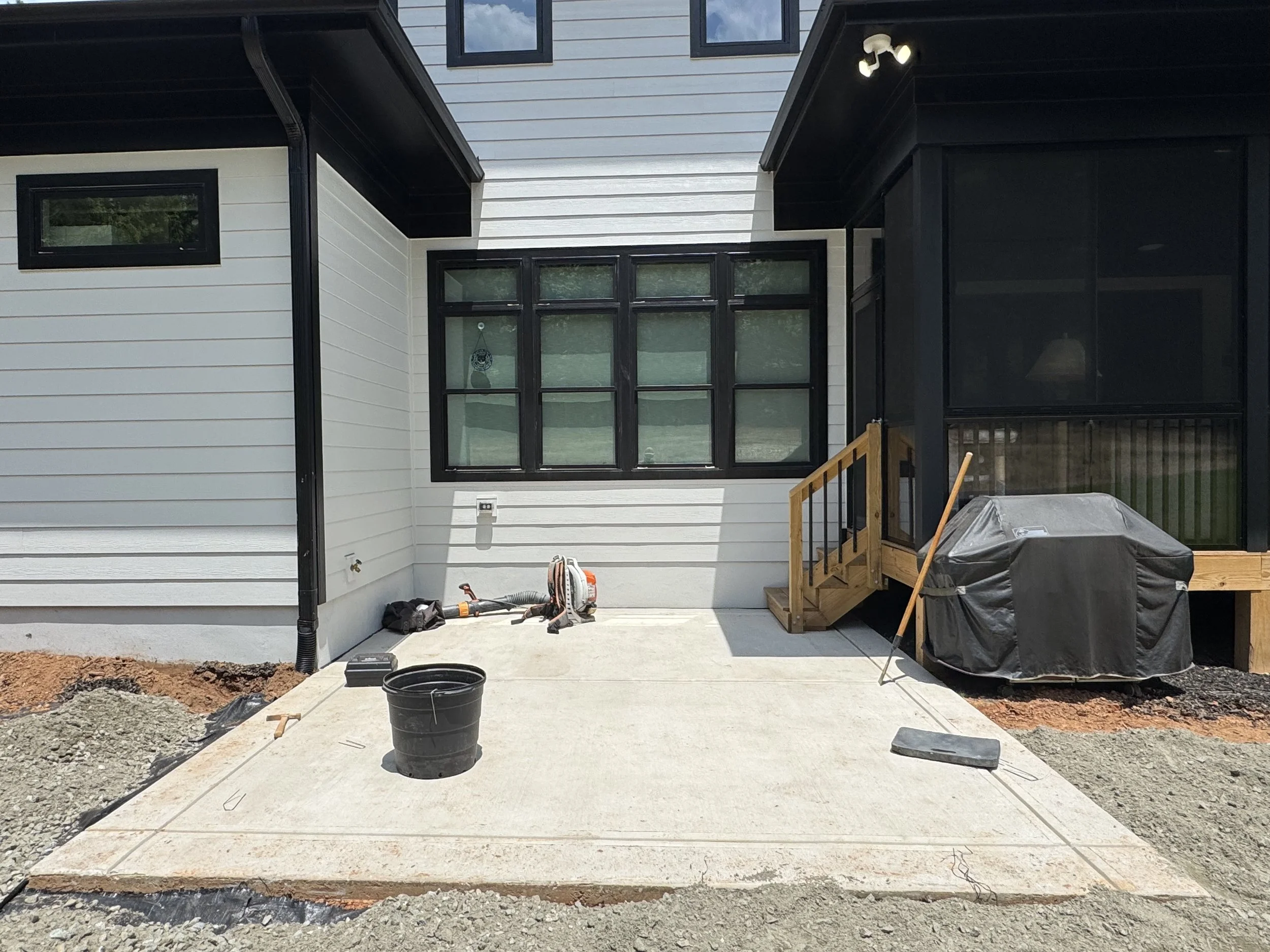The back of a house with white siding, black window frames, and black trim. There is a small staircase leading to a back porch or deck, covered with black railing. There are construction tools and materials, including a black bucket, a power tool, an