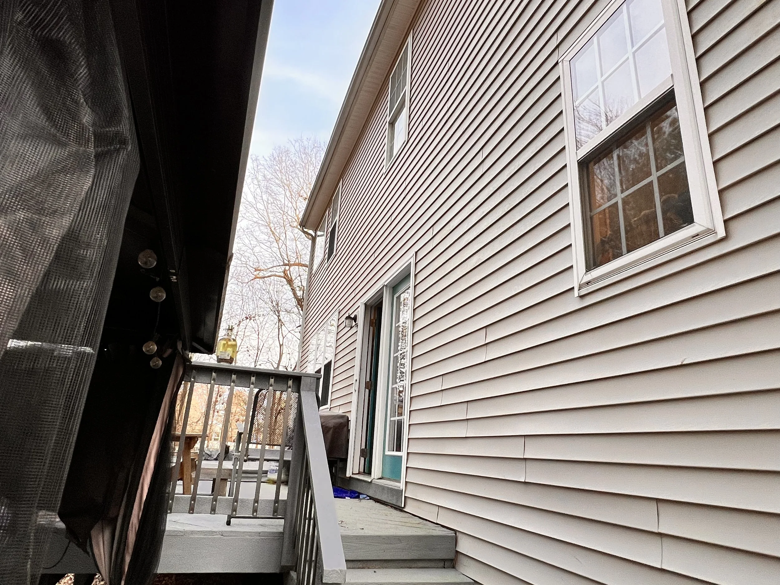 A view of a house exterior from a porch, showing beige horizontal siding, a sliding door, and a small deck with steps leading down.